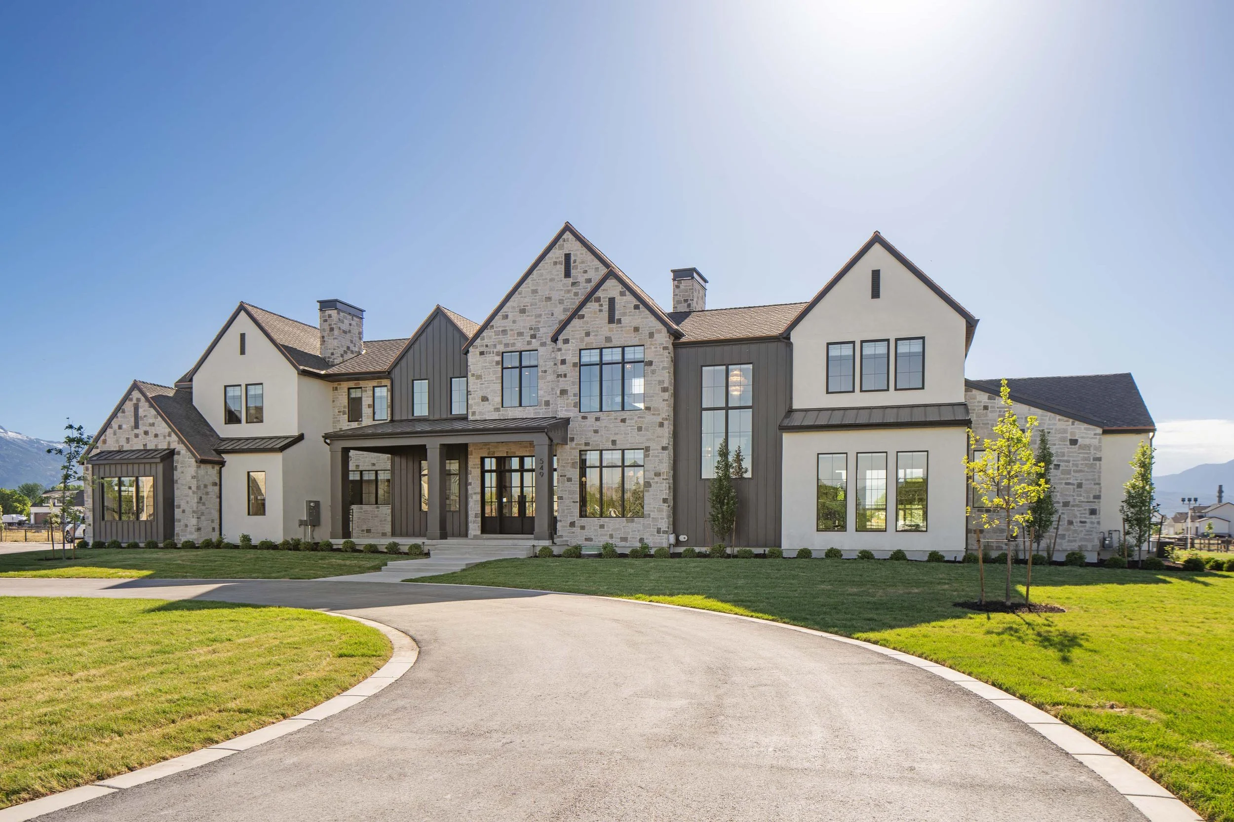Modern two-story house with stone and white exterior, large windows, and a curved driveway leading to the entrance, with a small lawn and young trees in the yard.