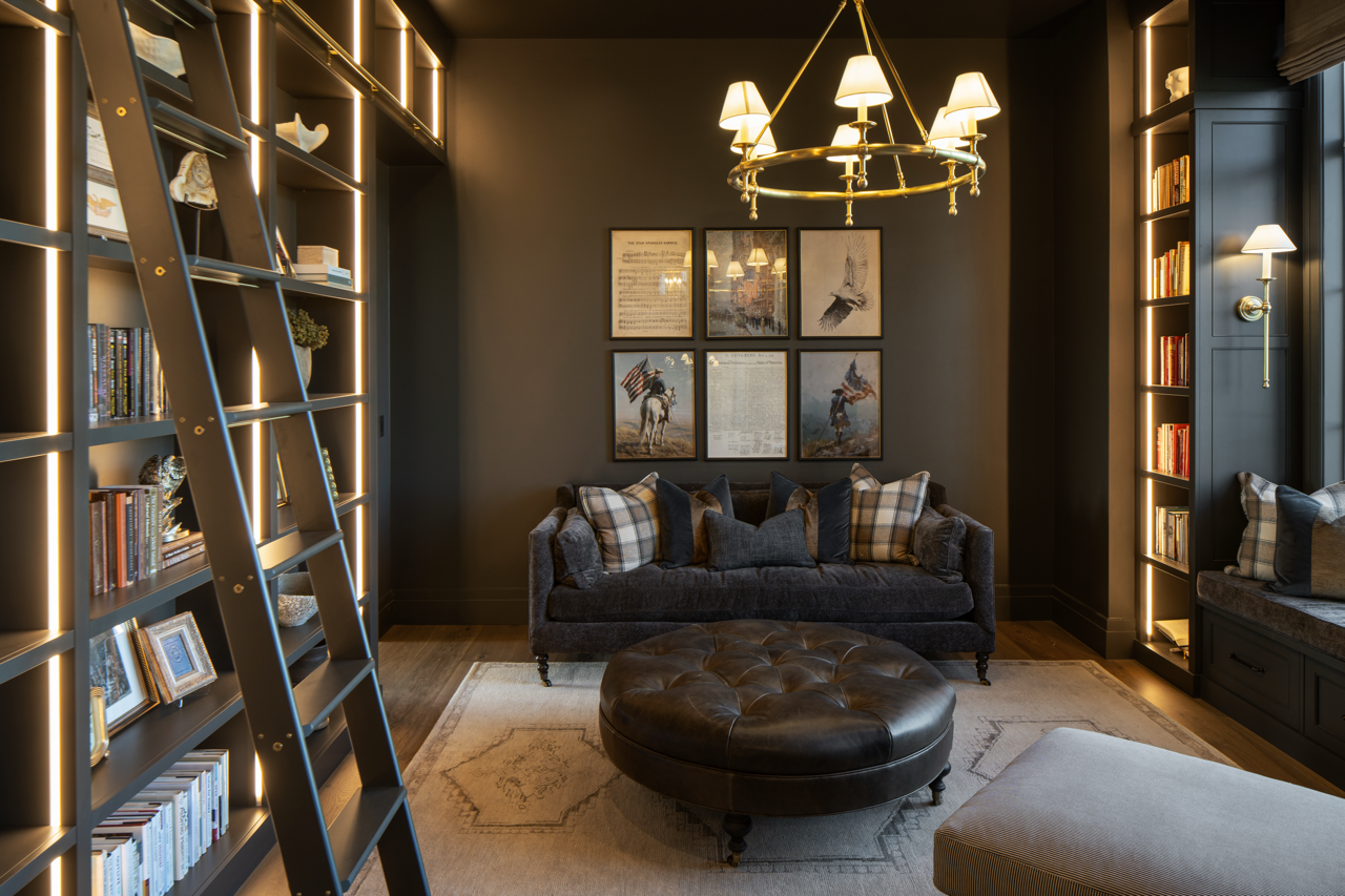 A cozy living room with dark walls, a dark sofa with plaid and solid pillows, a tufted leather ottoman, bookshelves with LED lighting, a ladder, framed art on the wall, a chandelier, and window seating.
