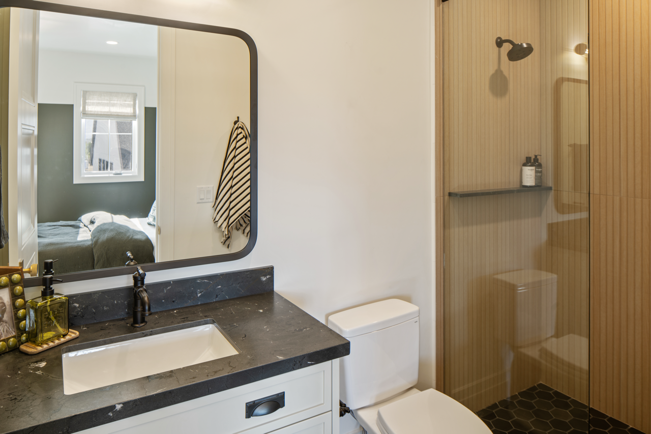 A modern bathroom with a black marble countertop sink, a large mirror, a white toilet, and a walk-in shower with wooden panel walls and black hexagonal tiles on the floor.