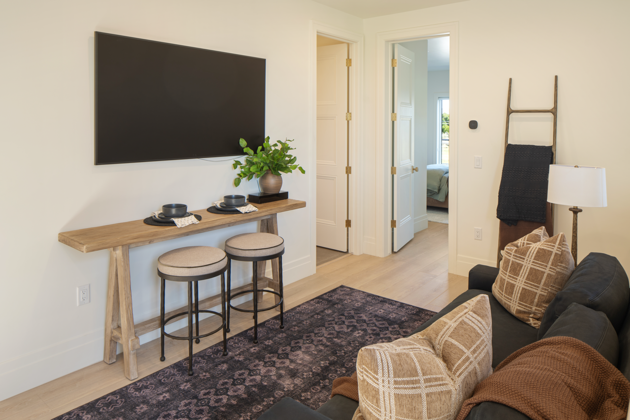 Living room with a black sofa, plaid pillows, a floor lamp, wall art in the shape of a ladder with a black blanket, a wall-mounted flat-screen TV, a wooden console table with two stools, a potted plant, and doors leading to other rooms.