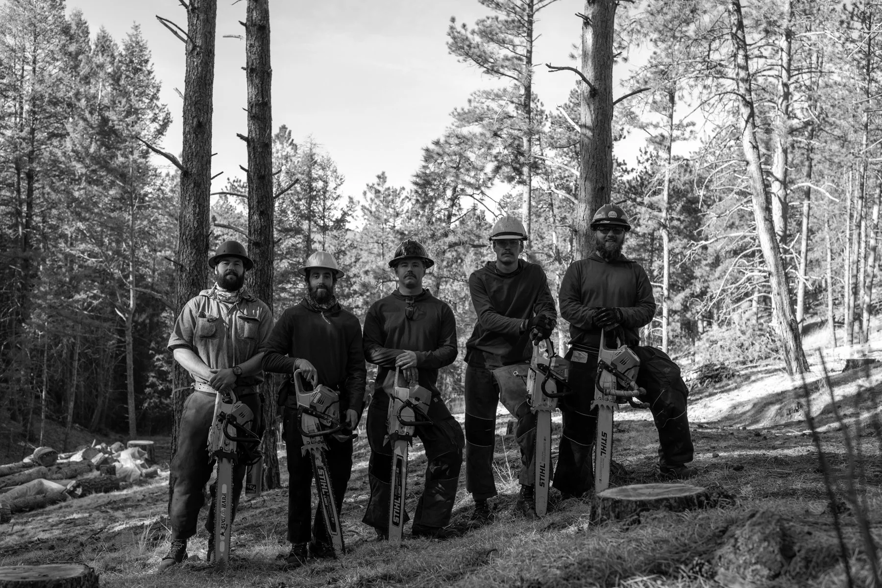 Five men standing in a forest, holding chainsaws, wearing safety helmets and gloves, with trees and chopped wood around them. The Forestwerx team