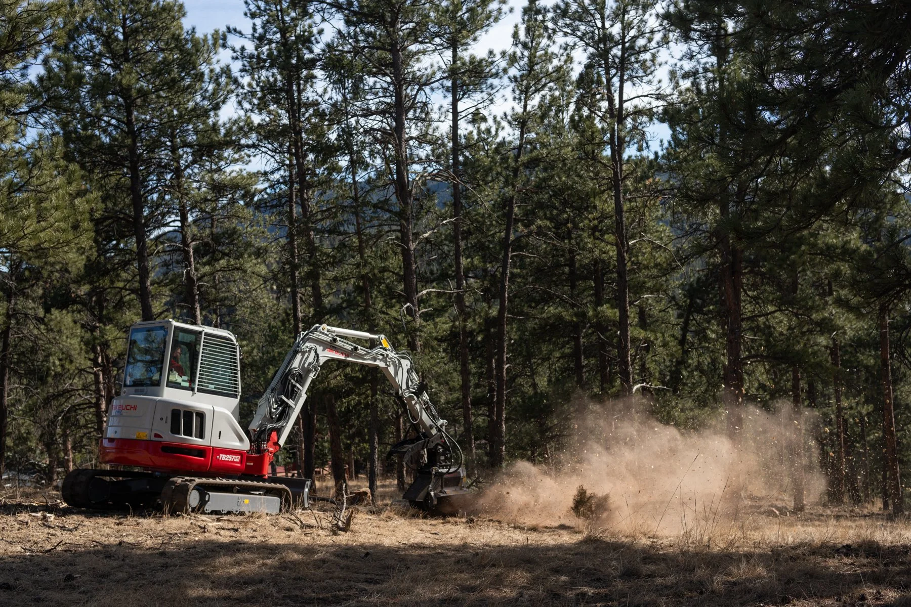 A Forestwerx excavator working in a forest clearing with tall pine trees in the background, kicking up dust as it moves.