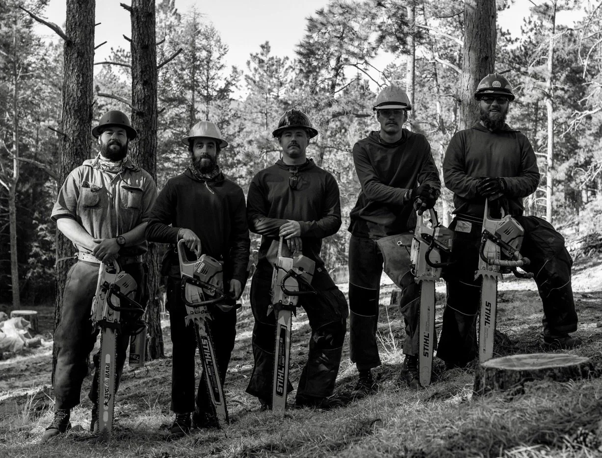 Five forestry workers standing outdoors in a forest, holding chainsaws, wearing protective helmets and gloves, in black and white.