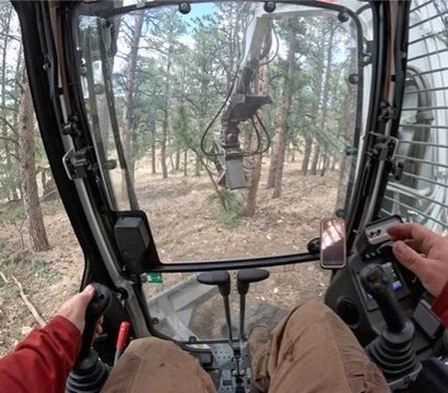 View from inside a construction vehicle cabin showing the operator's hands, control panel, and a forest with trees outside.