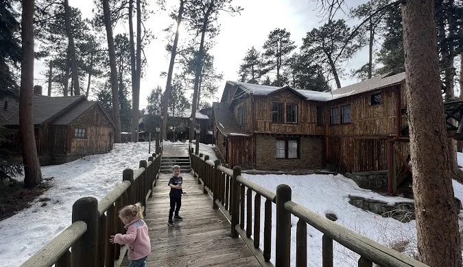 Two children playing on a wooden bridge in a snowy forest with wooden cabins in the background.
