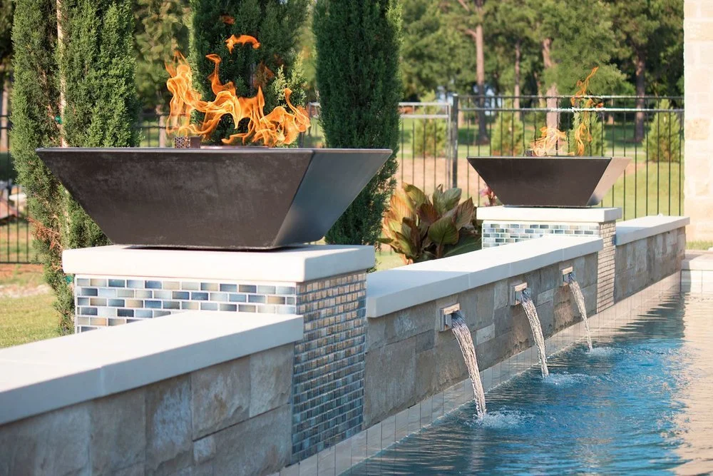 Outdoor water feature with two black fire bowls and cascading water spouts on a stone wall, surrounded by greenery.