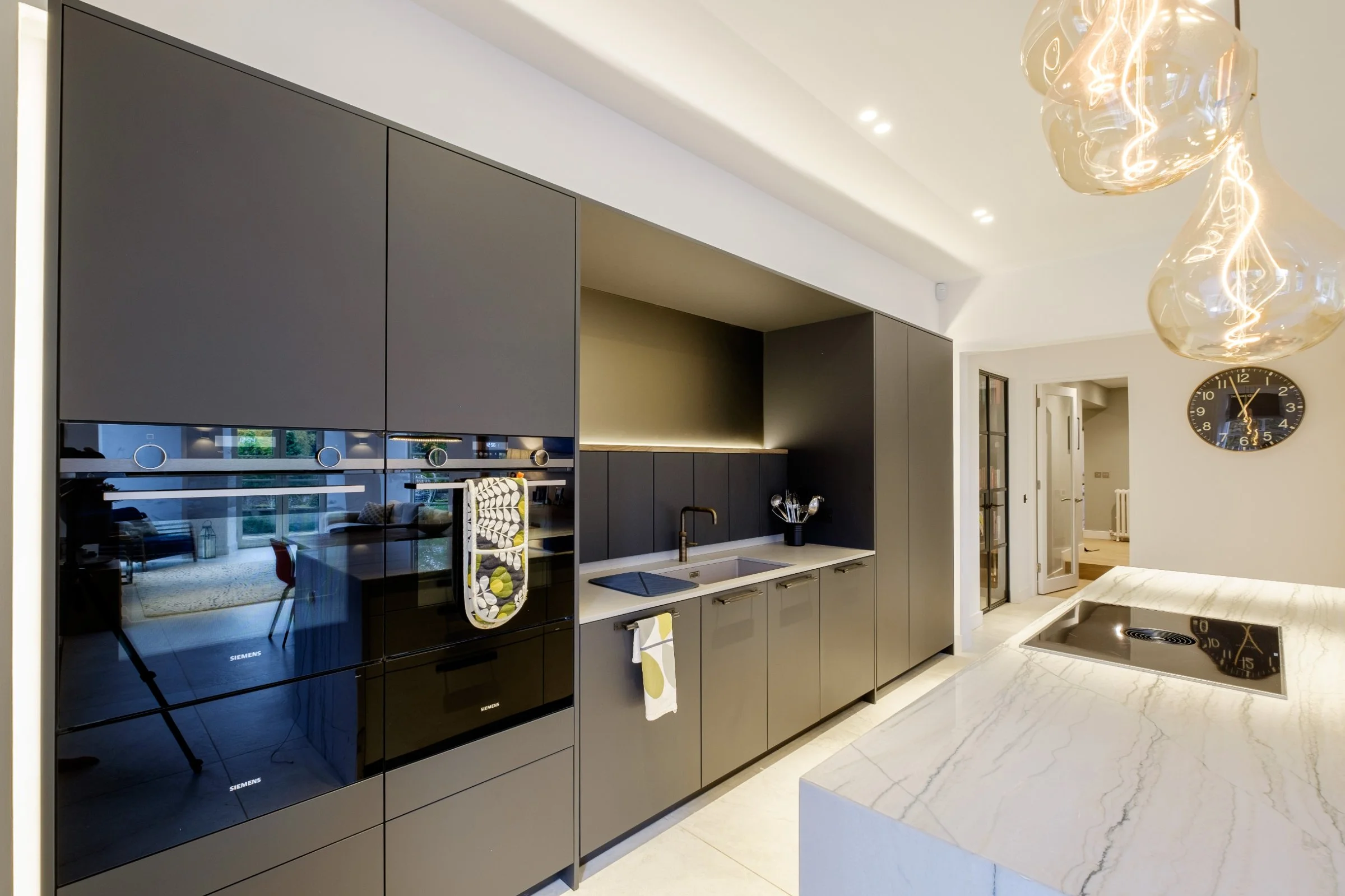 Modern kitchen with black cabinets, built-in appliances, marble countertop, and pendant lighting. A wall clock is displayed in the background.