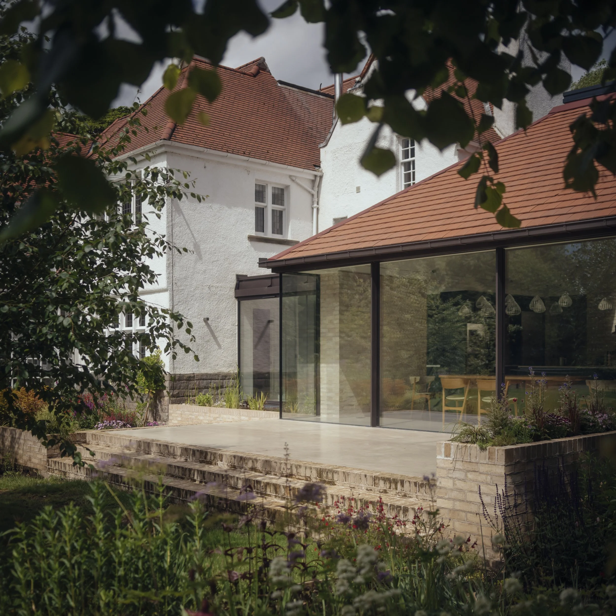 Modern house extension with glass walls, brown tiled roof, and a brick patio surrounded by greenery.