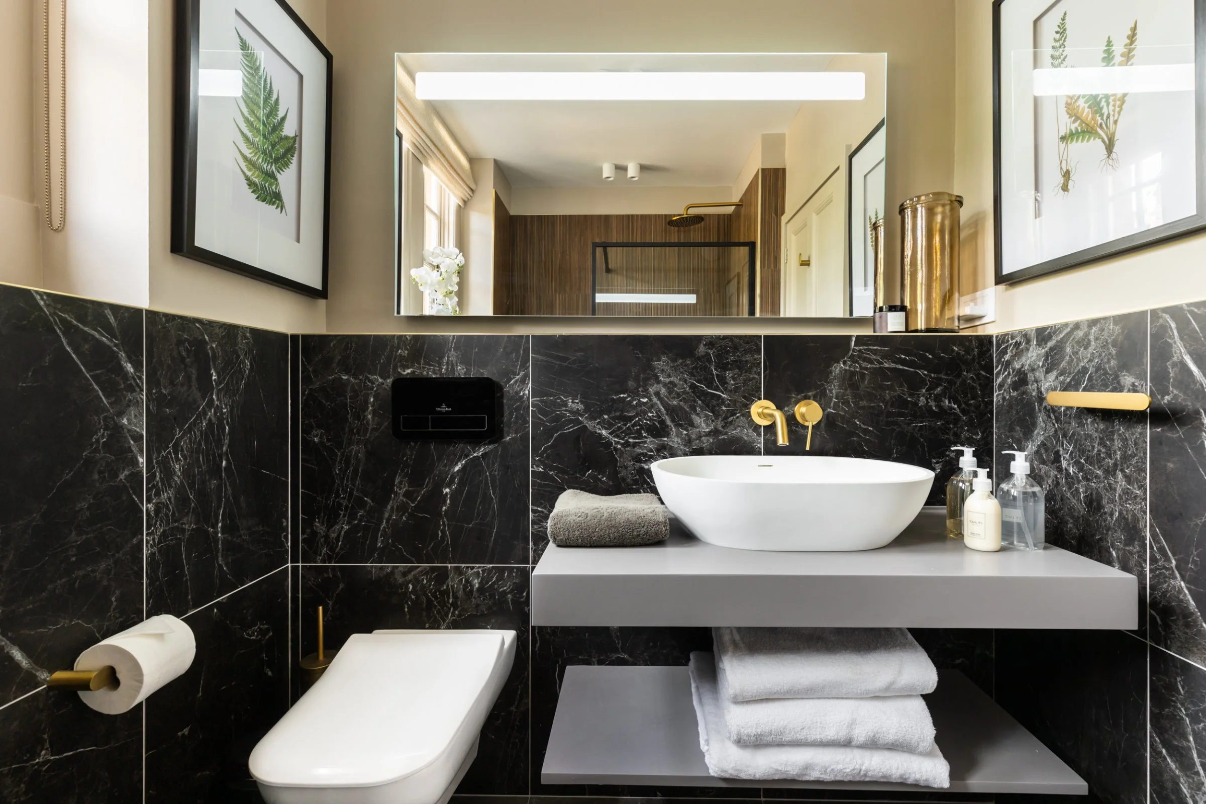 Modern bathroom with black marble walls, gold faucet, white vessel sink, and toilet; framed fern artwork on walls, mirror reflecting shower area, and toiletries with towels arranged neatly.