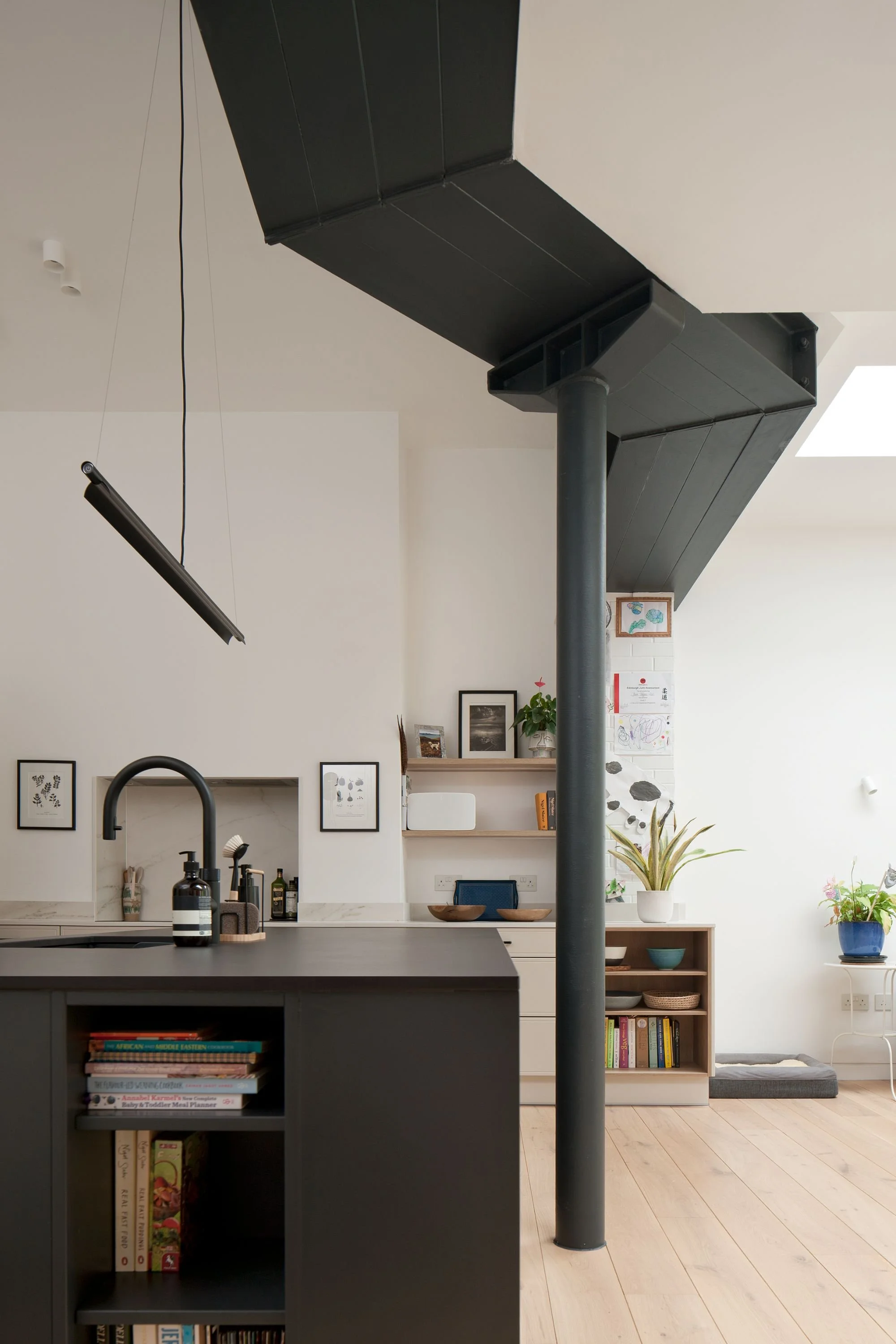 Modern kitchen with a black island, wooden shelves, and plants. The room features a unique black overhead structure and minimalist decor, including books and framed pictures on the wall.