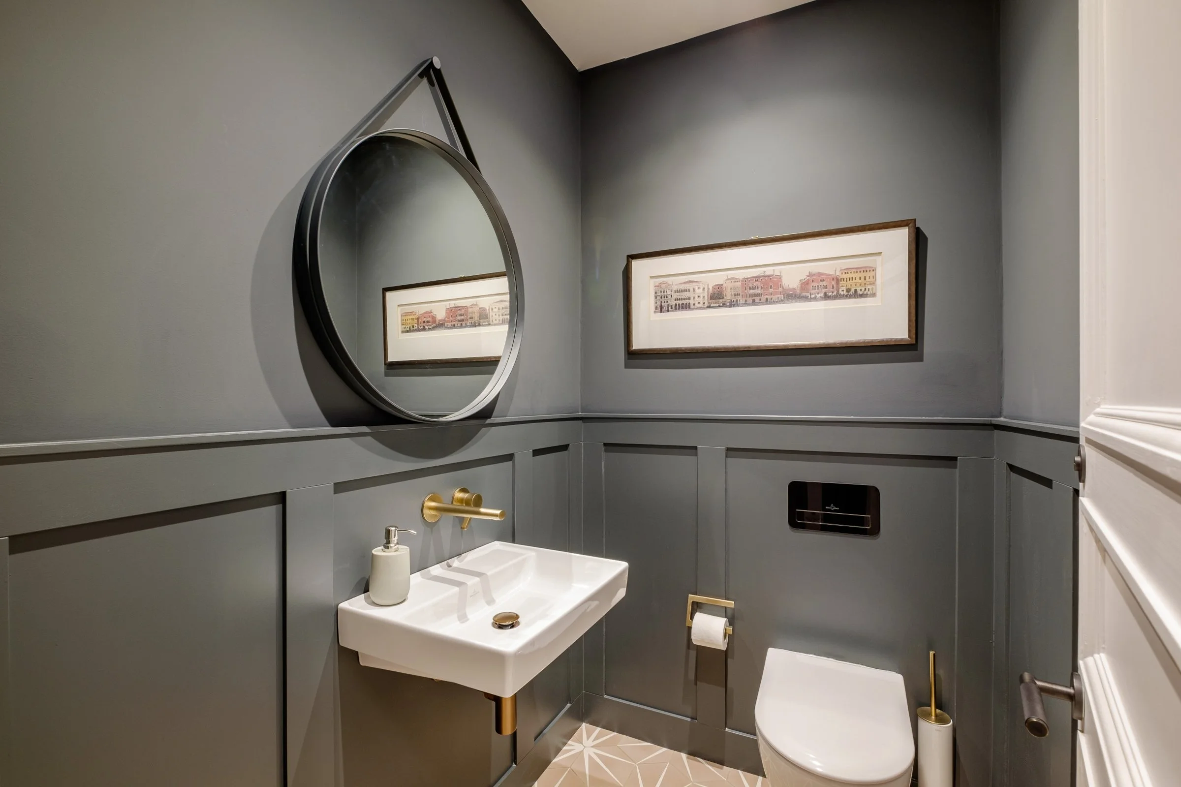 Modern bathroom interior with dark walls, a round mirror, wall-mounted sink with a gold faucet, framed artwork, and a toilet with a geometric floor tile pattern.