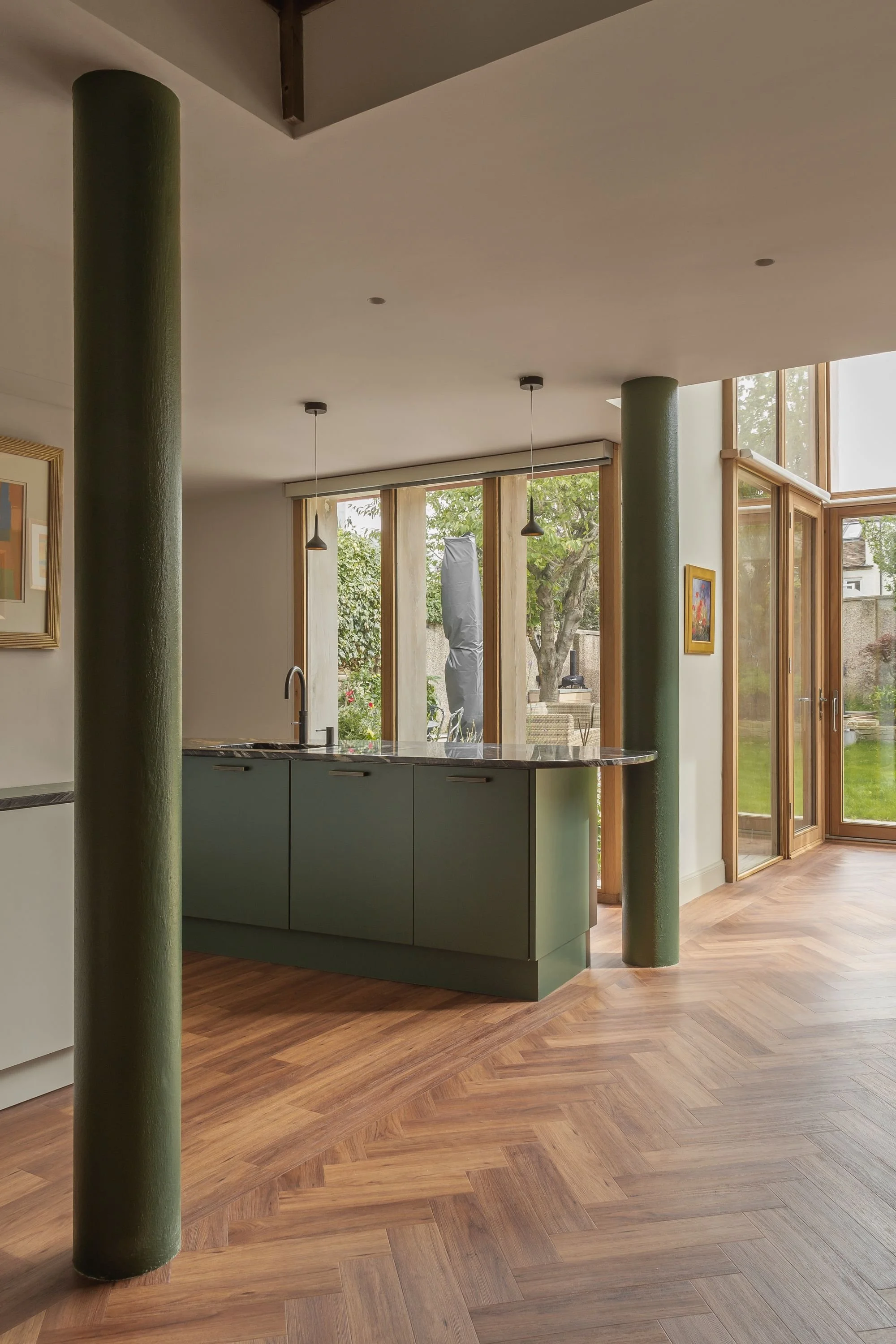 Modern kitchen with green cabinets, wooden floors, large windows, and green columns, overlooking a garden.