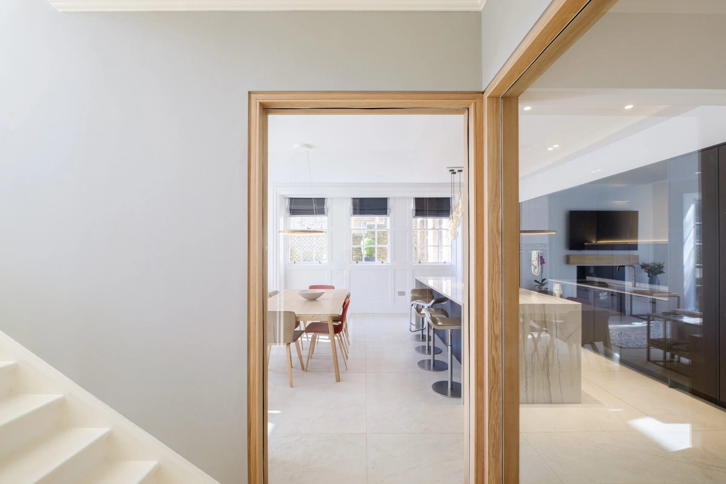 Modern dining area with a wooden table, assorted chairs, and a kitchen island with bar stools, large windows, and pendant lights.