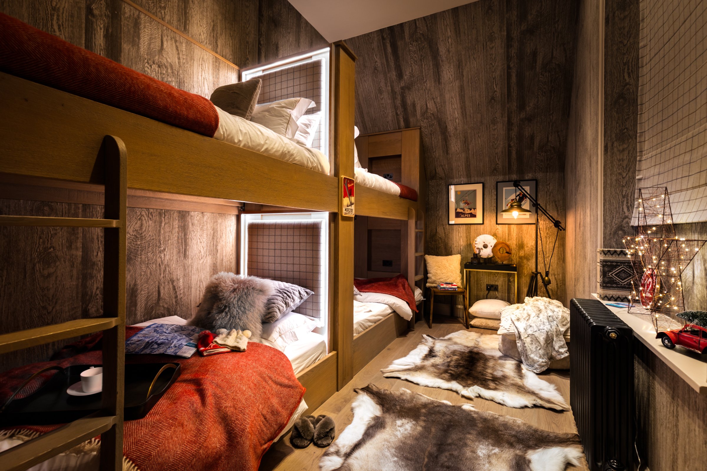 Cozy small room with wooden bunk beds, soft lighting, and warm decor. Includes fur rugs, a padded armchair, and decorative lights. Room features framed art on wood-panel walls and a window with a grid pattern shade.