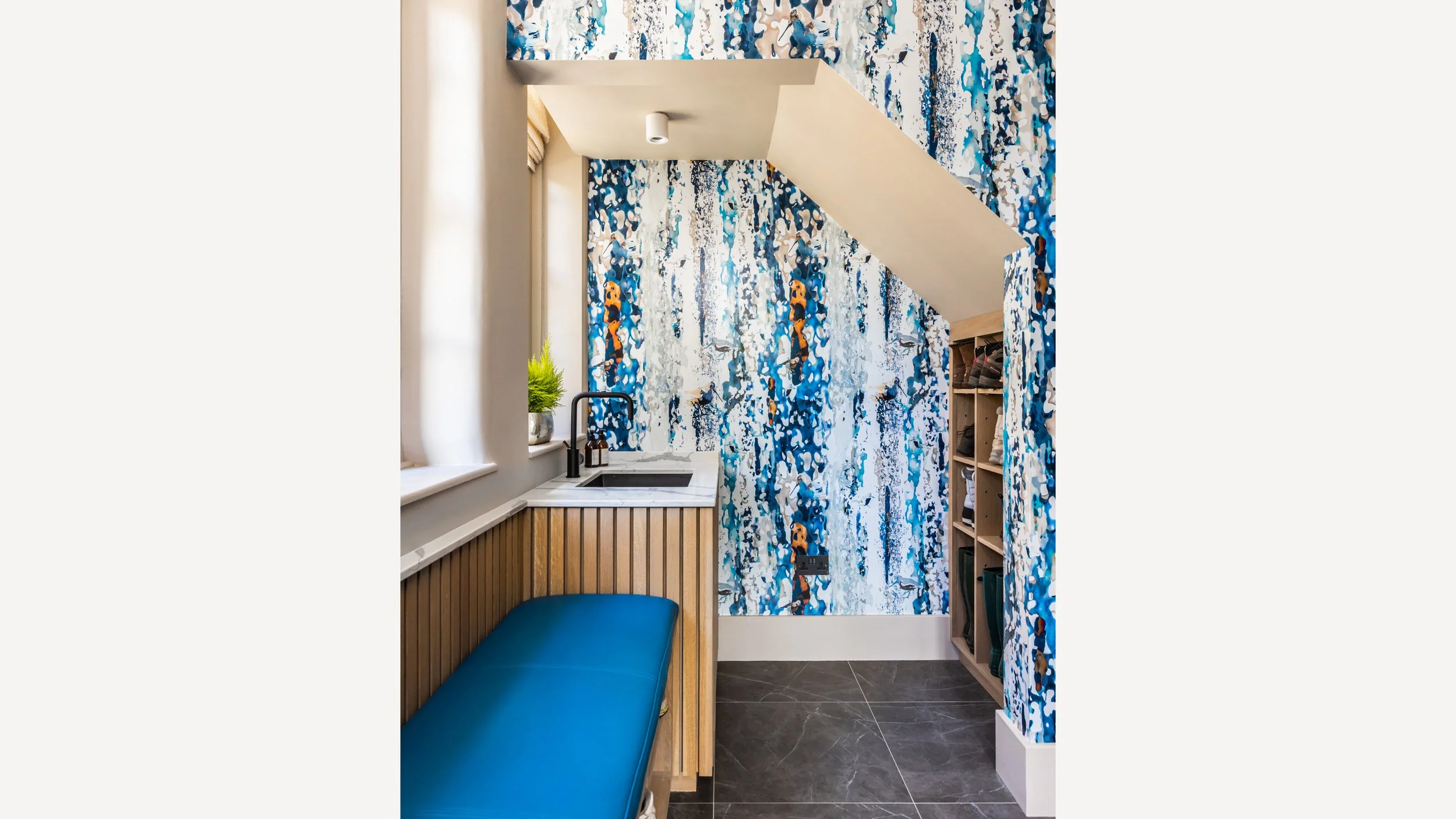 Small room with blue abstract patterned wallpaper, a bench with a blue cushion, a countertop with a sink and faucet, shelving unit, and a small plant on the windowsill.