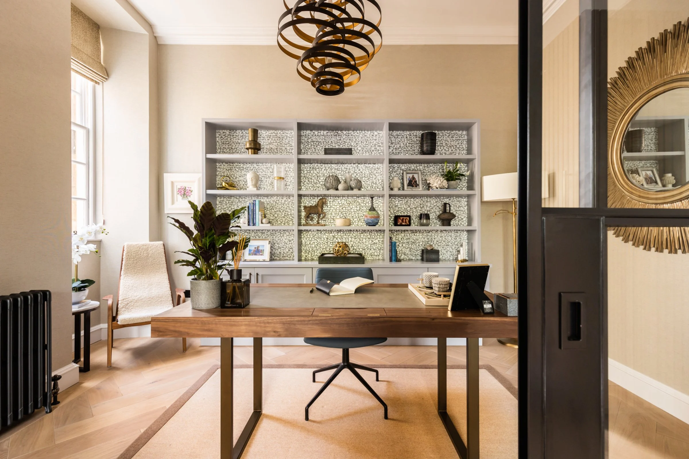 Modern home office with a wooden desk, black chair, shelves with decorative items, indoor plants, large window, and an ornate ceiling light fixture.