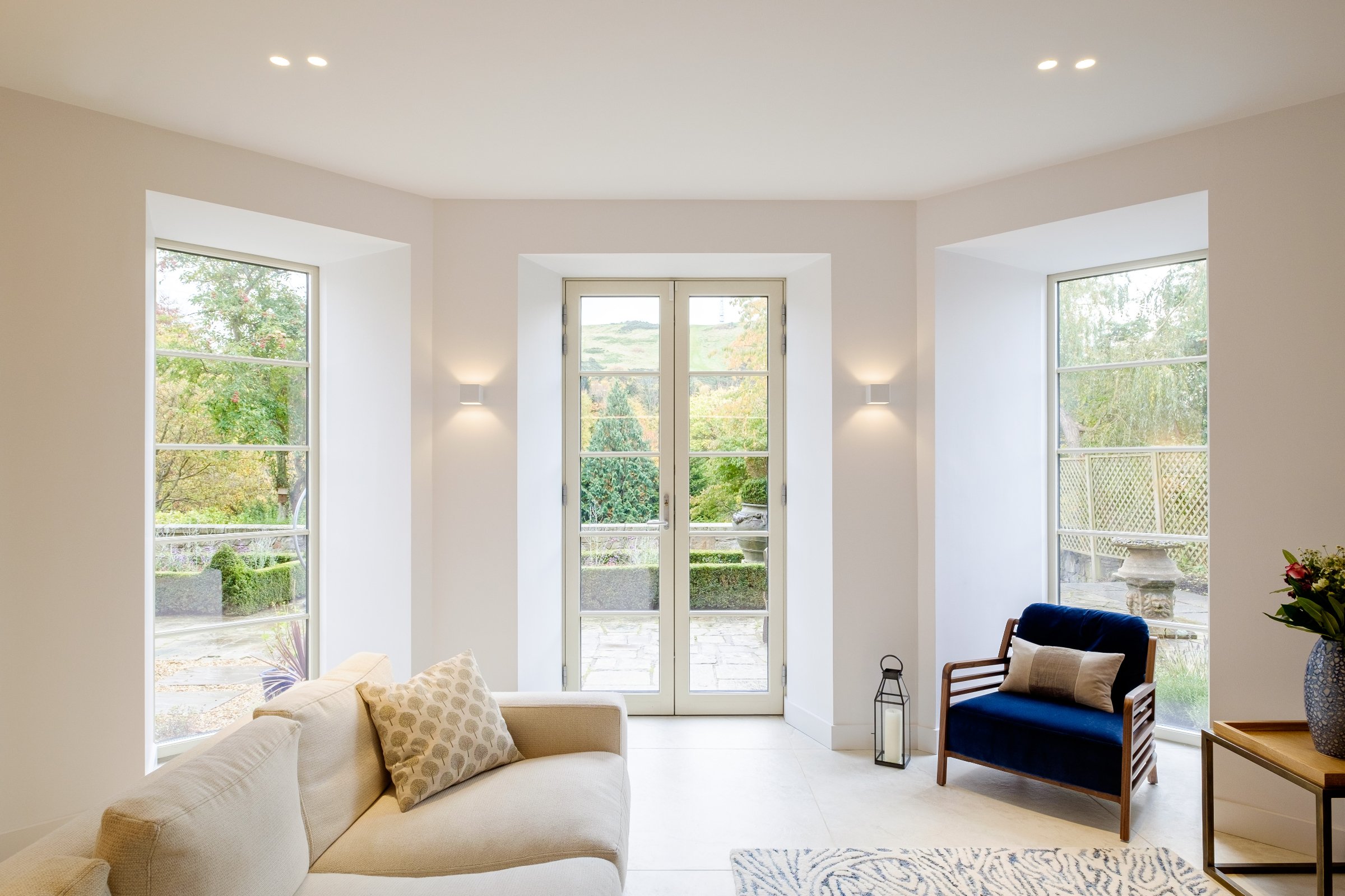 Modern living room with large windows, white walls, a beige sectional sofa, a blue armchair, patterned rug, indoor plants, and outdoor view.