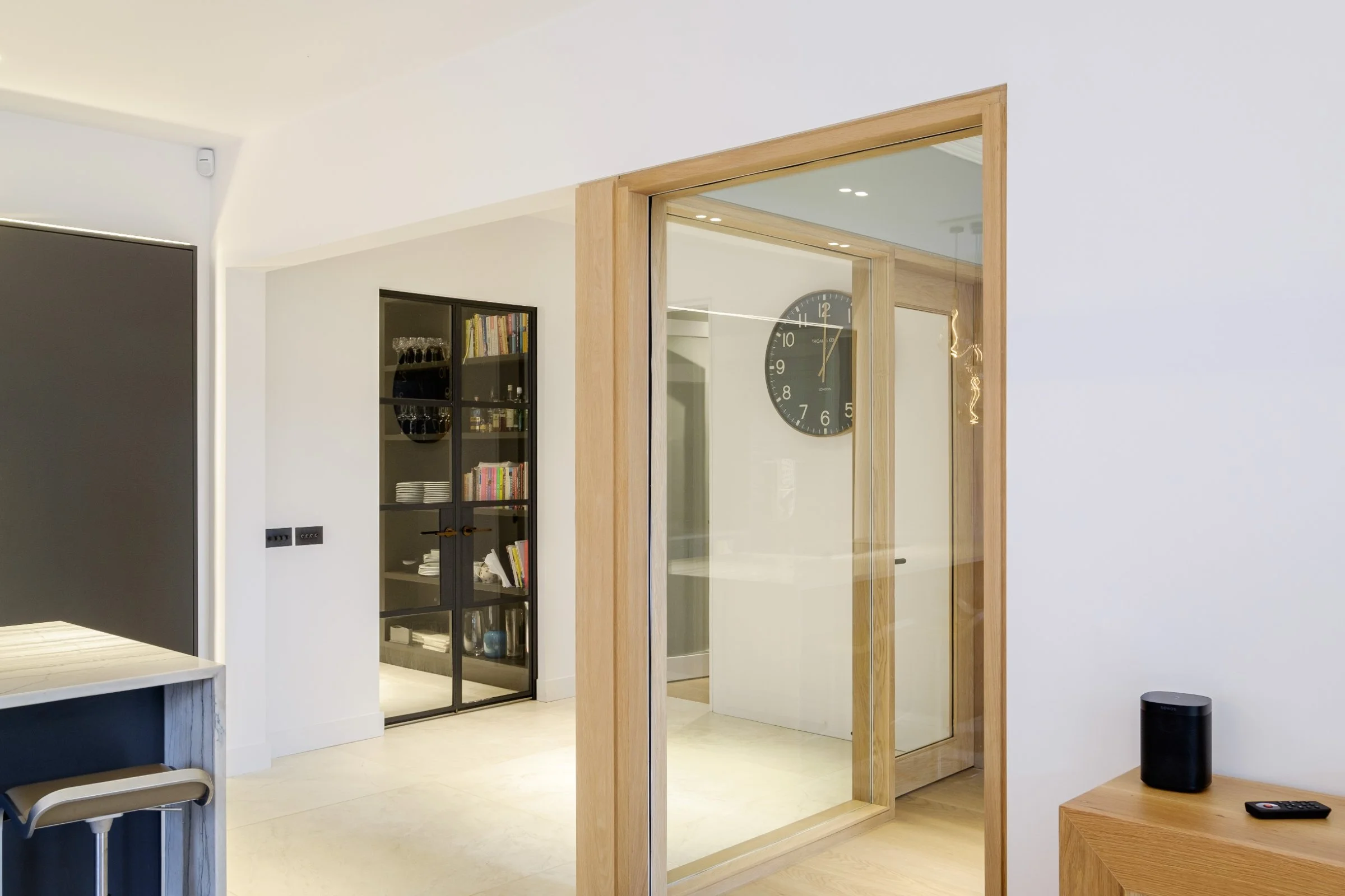 Modern interior with glass doors, a wall clock, shelving with books and dishes, a bar stool, and a small black speaker on a wooden cabinet.
