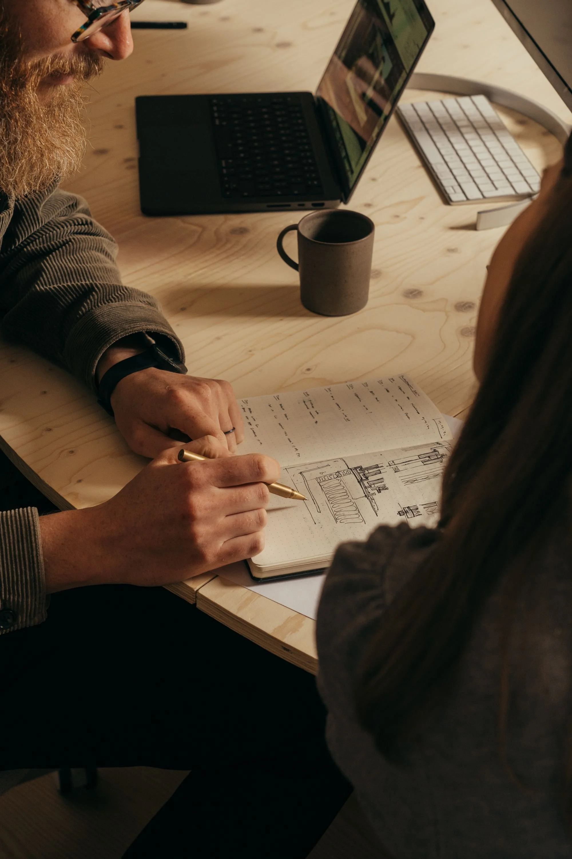 A person with glasses and a beard is drawing on a sketchbook at a wooden table, with a pen in hand. There is a black coffee mug, a laptop, and a keyboard on the table.