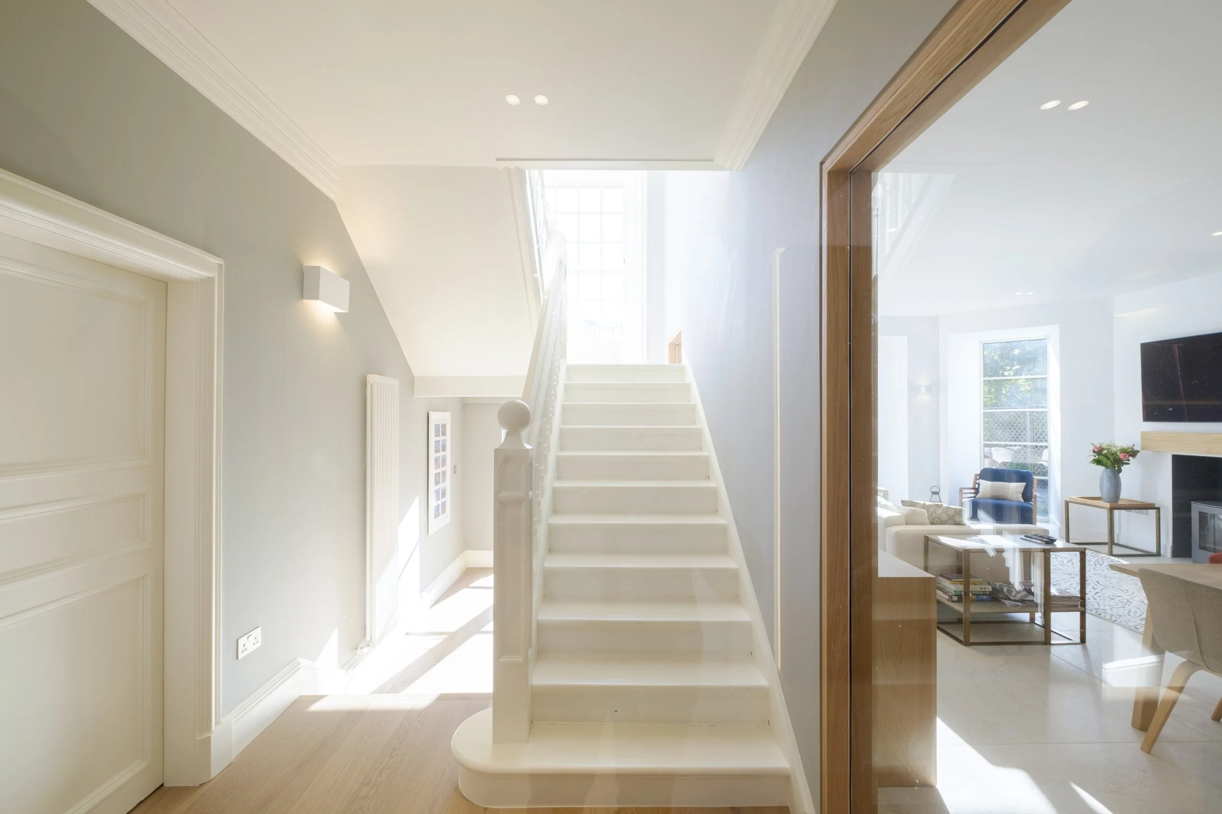 Bright interior hallway with white staircase, leading up to a window. Adjacent to a modern living room with wooden furniture and large windows.