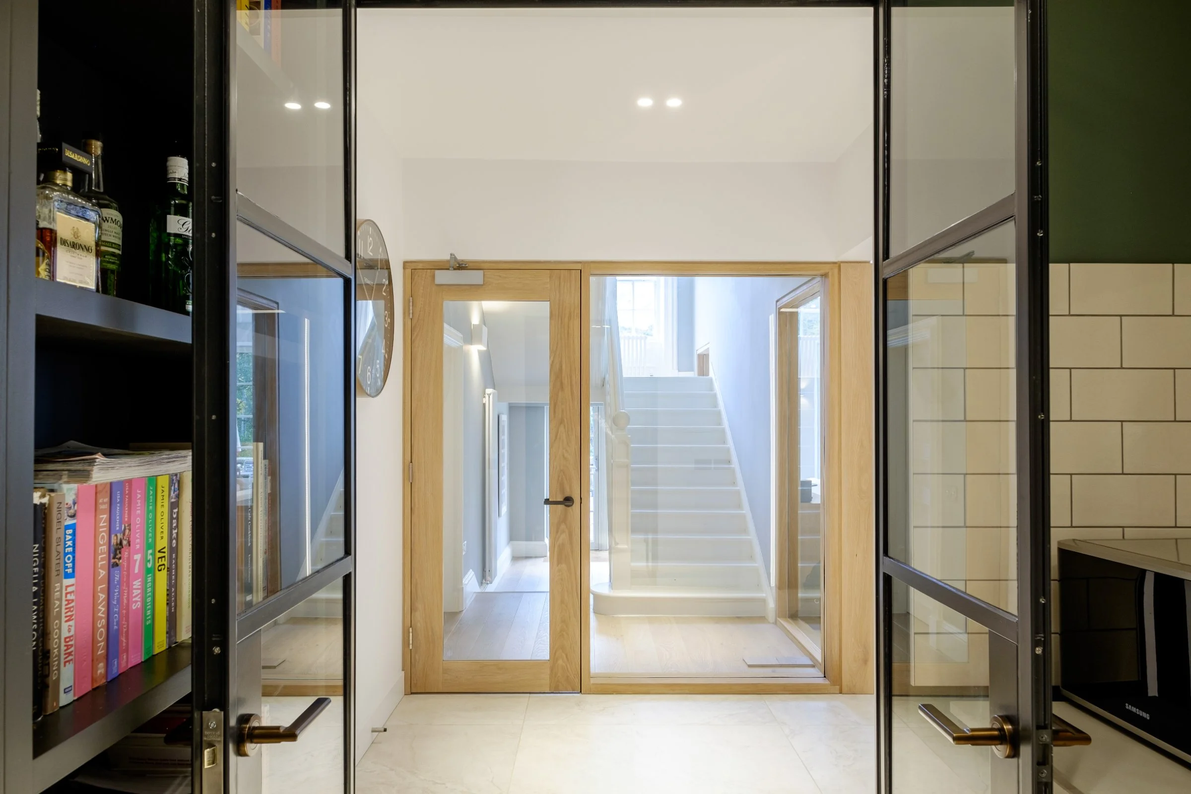 Modern interior hallway with wooden and glass doors, visible staircase, and shelving with books and bottles.