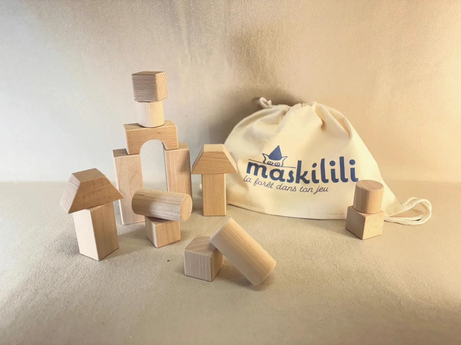 Différents blocs de bois formant des structures de maisons et une pochette en tissu avec le logo Maskilili, jouet en bois pour enfants.