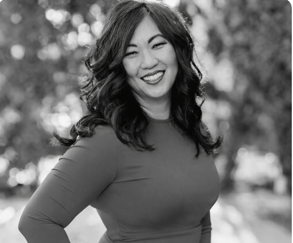 Black and white photo of a smiling woman with long curly hair, wearing a long-sleeve top, outdoors with blurred trees in background.