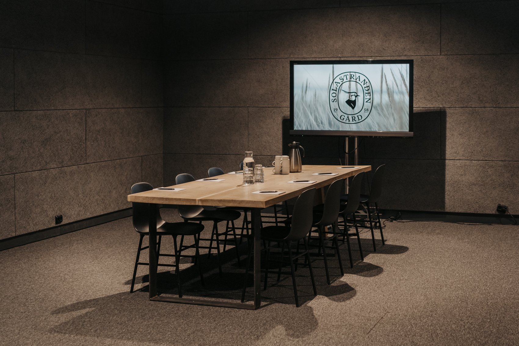 Møterom med lang konferansebord, stoler, serveringskanne og skjerm med logoen «Solastranden Gård».