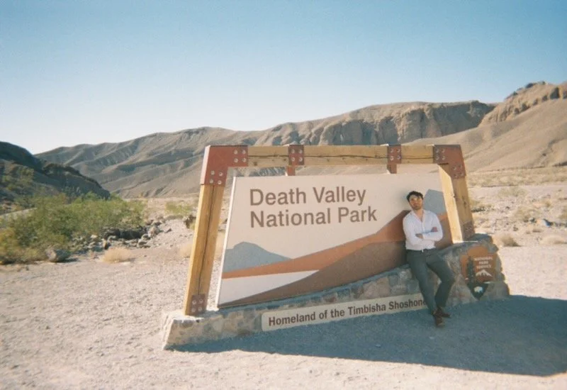 A person leaning against a large sign that reads 'Death Valley National Park' in a desert landscape with mountains in the background.
