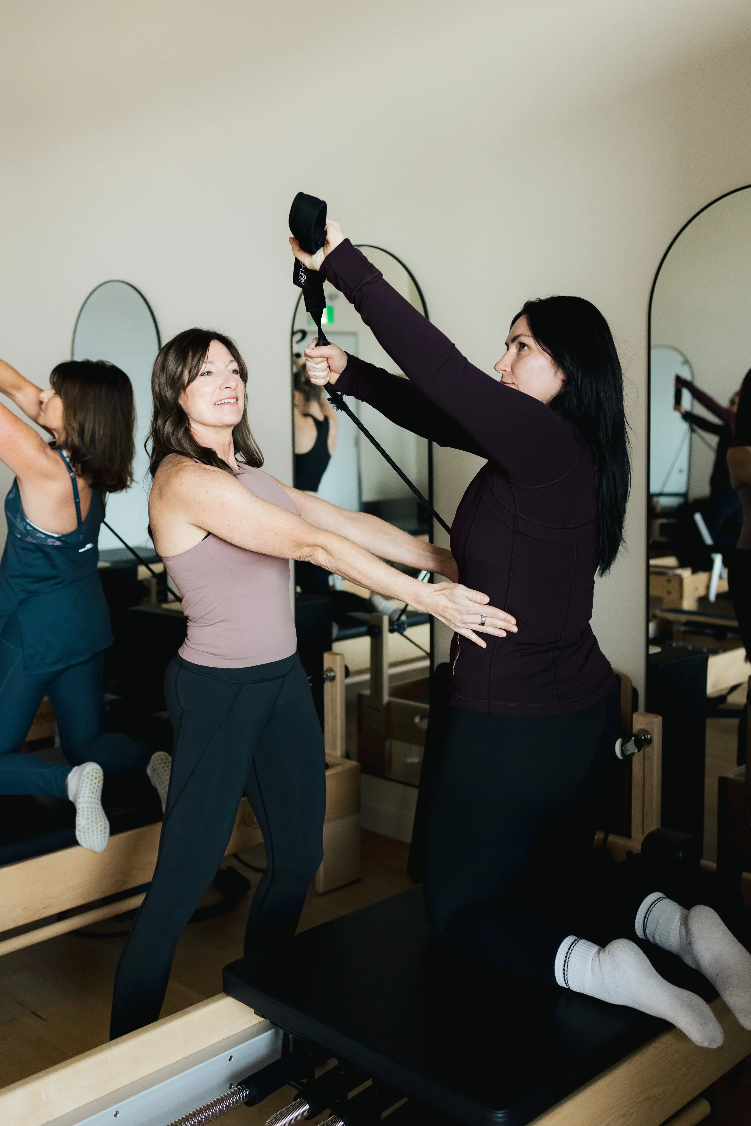 A woman with dark hair kneeling on a Pilates reformer machine during a workout class, being assisted by an instructor in a fitness studio.
