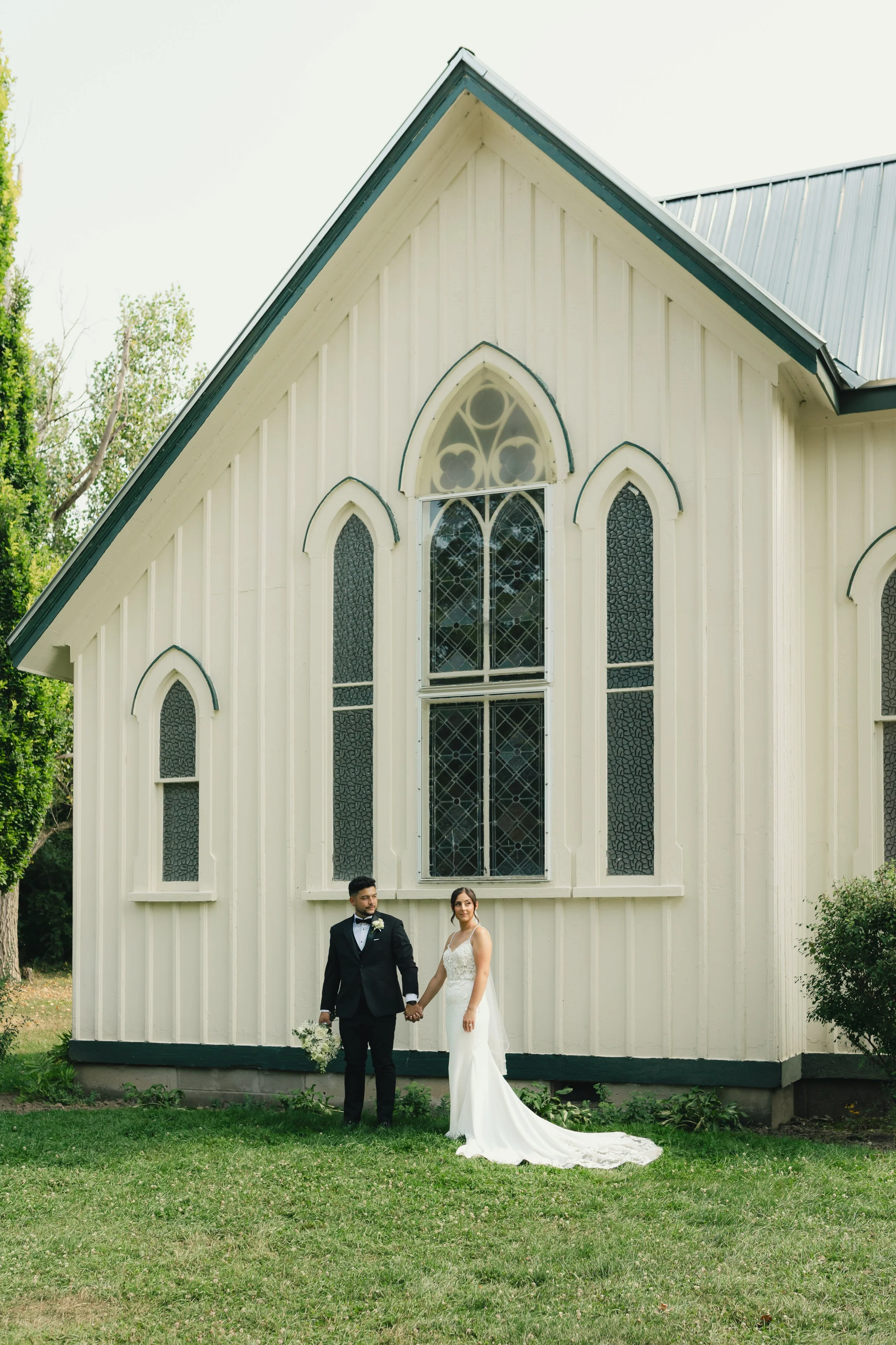A bride and groom holding hands in front of a white church with stained glass windows, dressed in wedding attire, outdoors on a grassy area.
