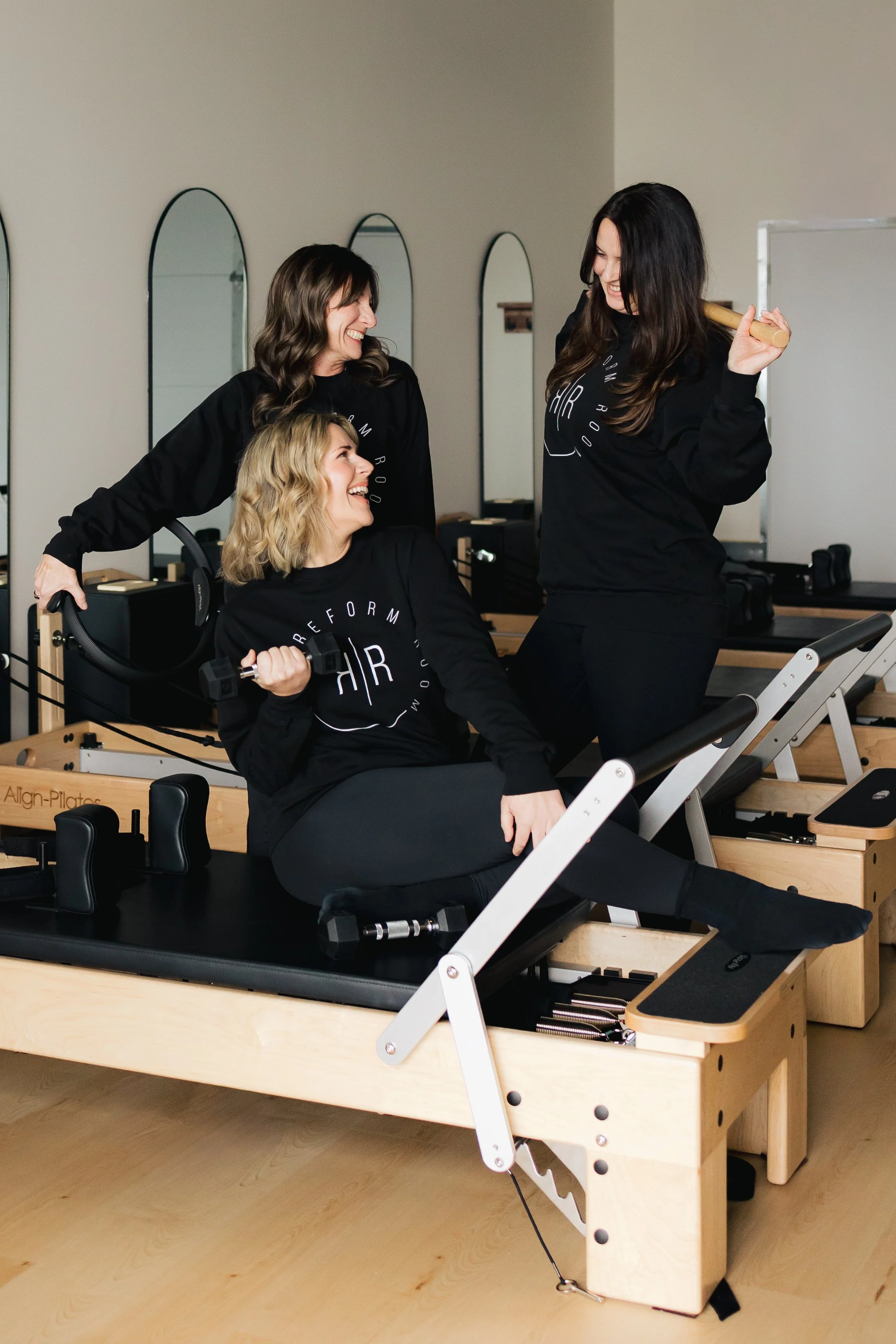 Three women in black workout apparel smiling and chatting in a Pilates studio, with one woman holding a dumbbell and another holding a wooden stick, surrounded by Pilates equipment and mirrors.