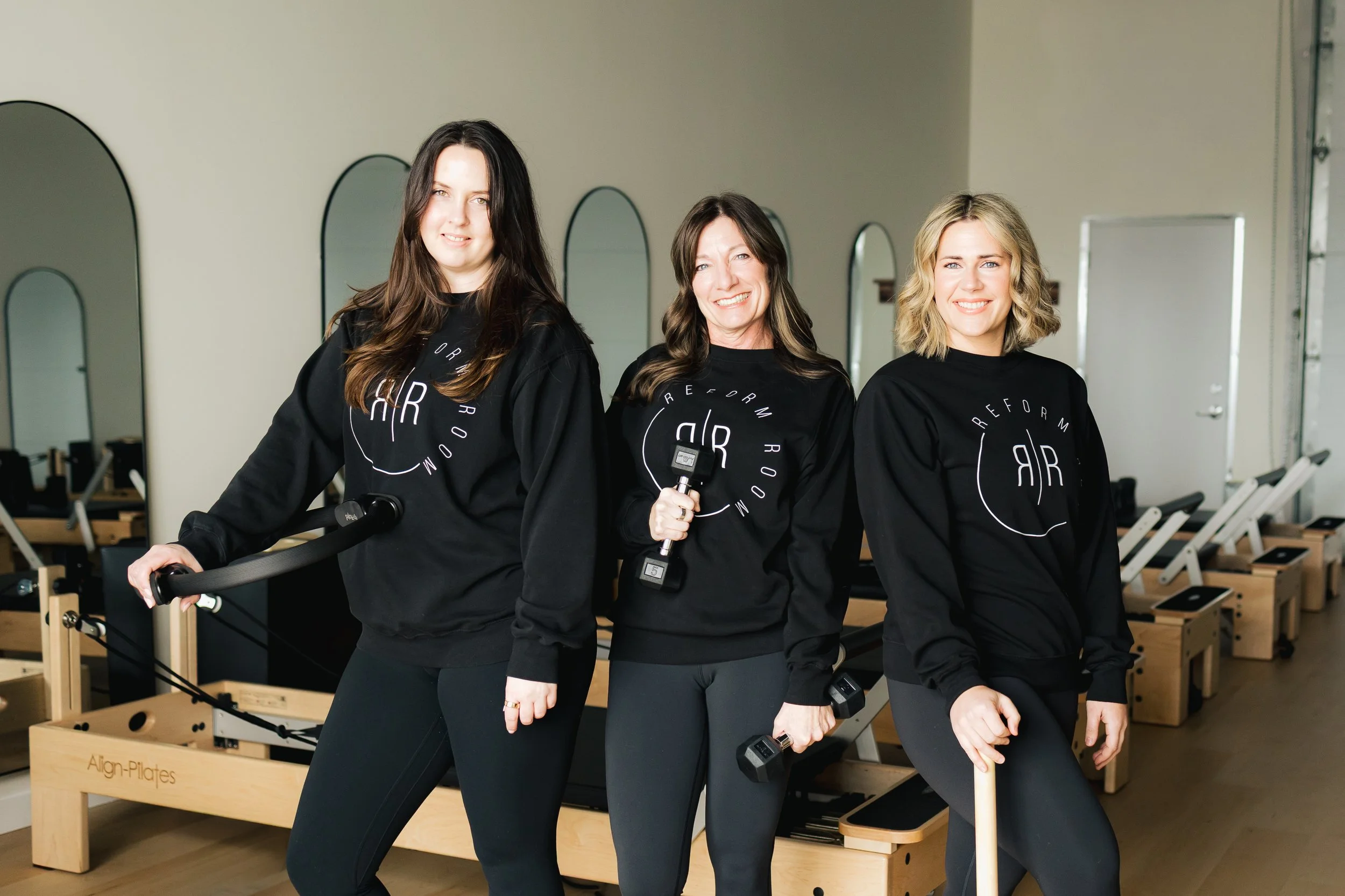Three women in workout attire holding dumbbells and a Pilates ring in a fitness studio with Pilates reformer machines in background.