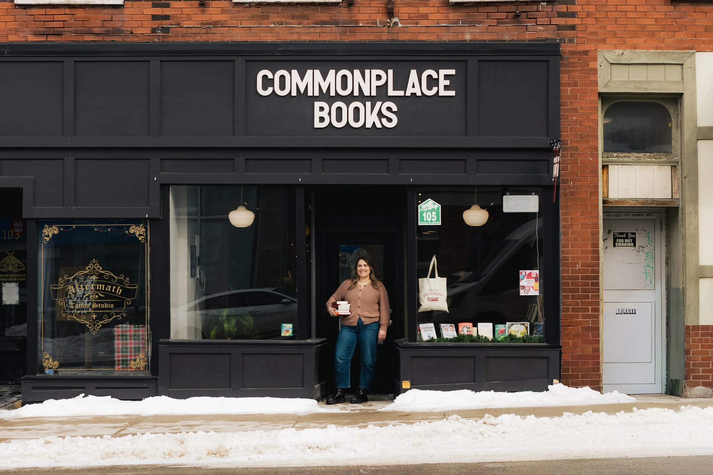 A woman standing outside a bookstore named Commonplace Books, holding a stack of books, with snow on the sidewalk in front.