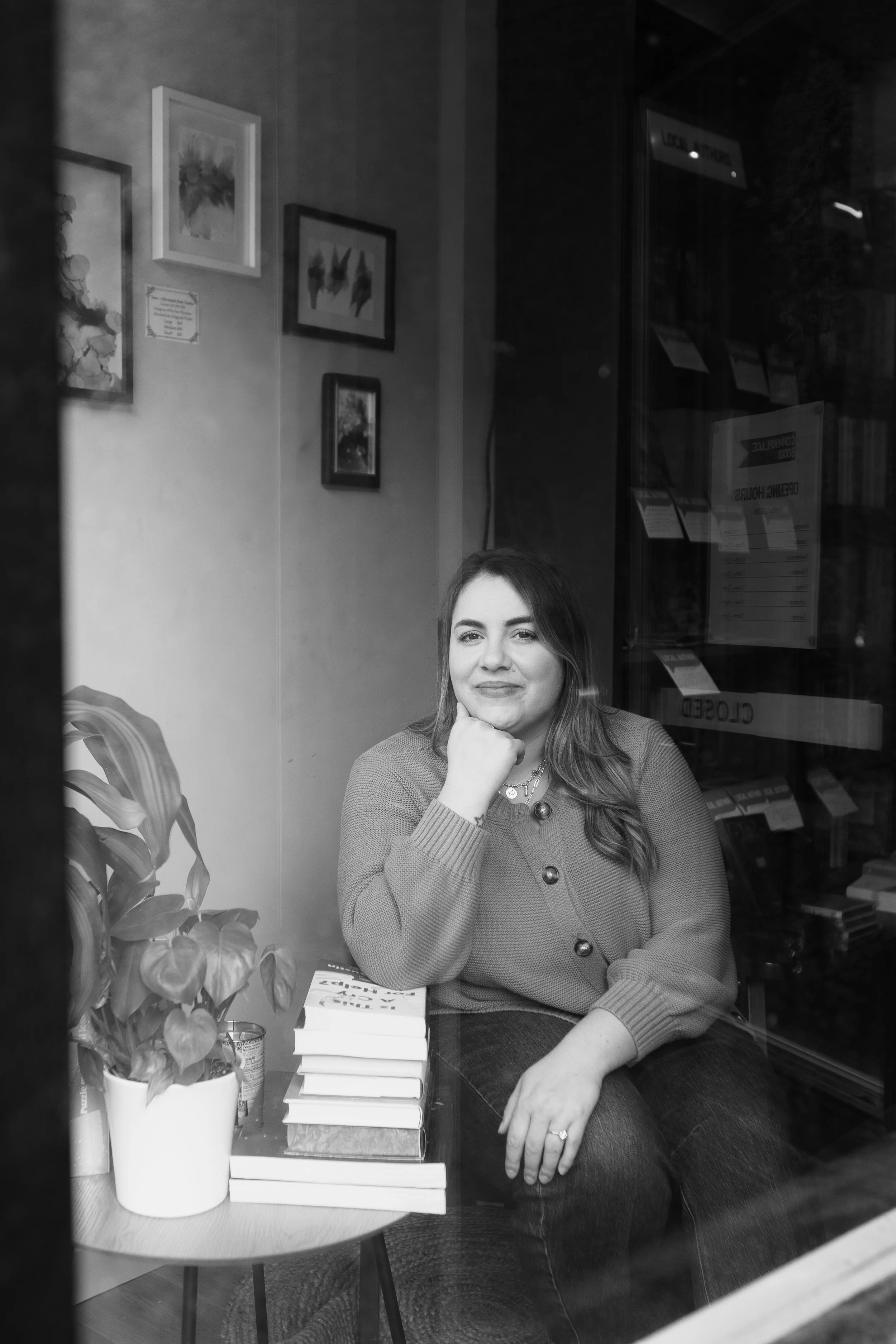 A woman sitting inside a bookstore, looking out the window, with a stack of books and a plant on a small table beside her.