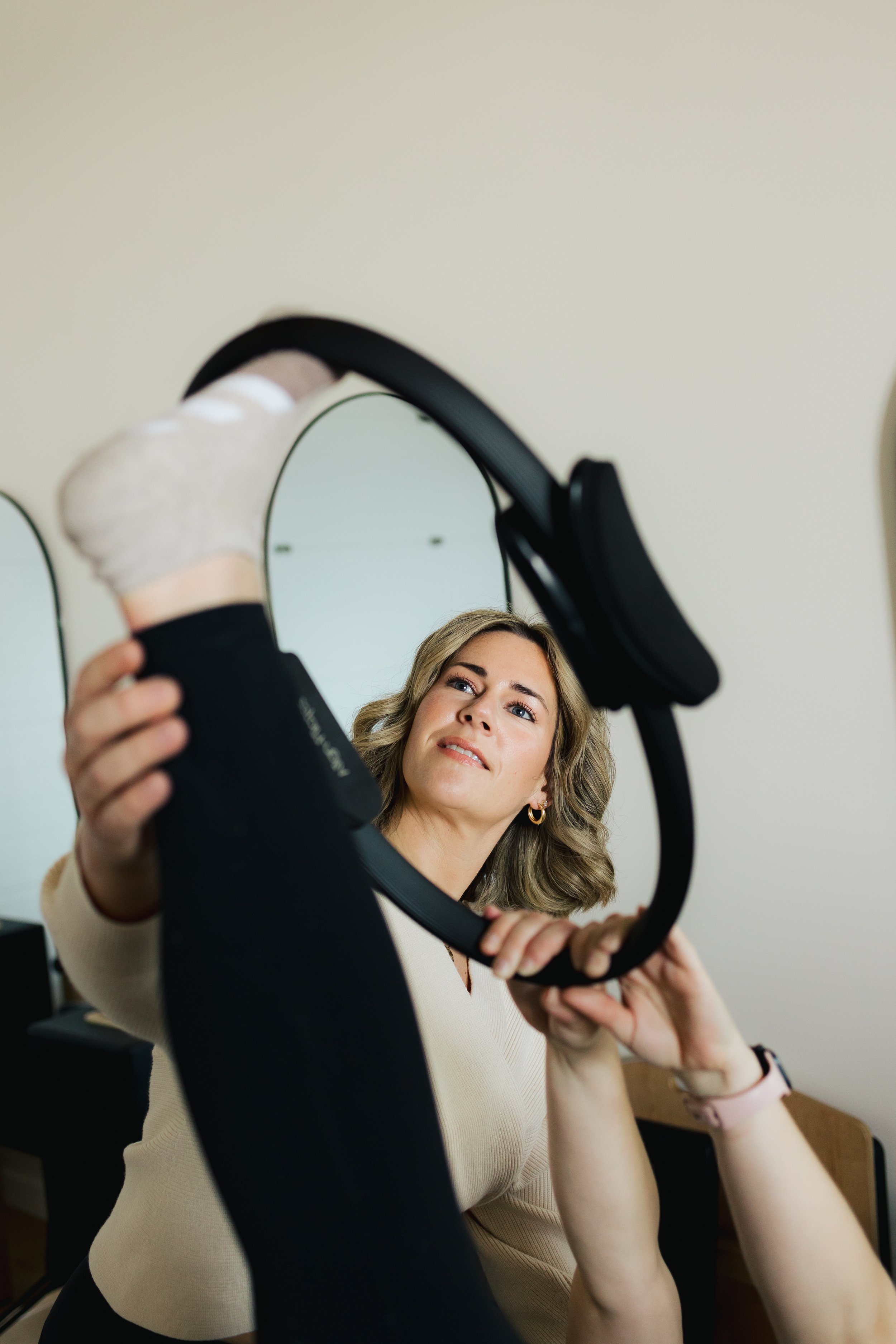 A woman with wavy blonde hair looking into a circular mirror while holding it up, with a neutral background and a piece of furniture visible in the room.