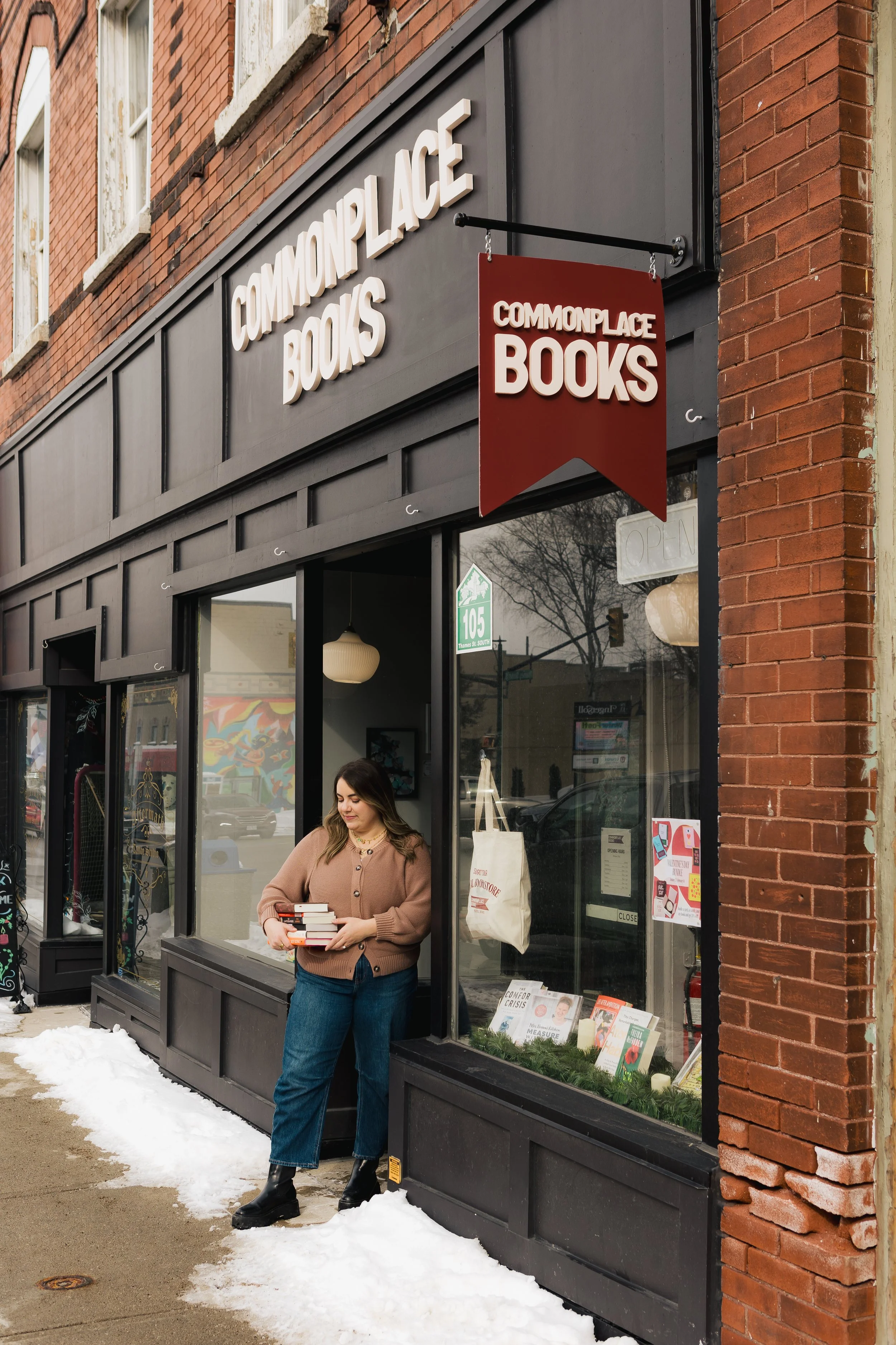 A woman standing outside a bookstore called Commonplace Books, holding a stack of books, with snow on the sidewalk.