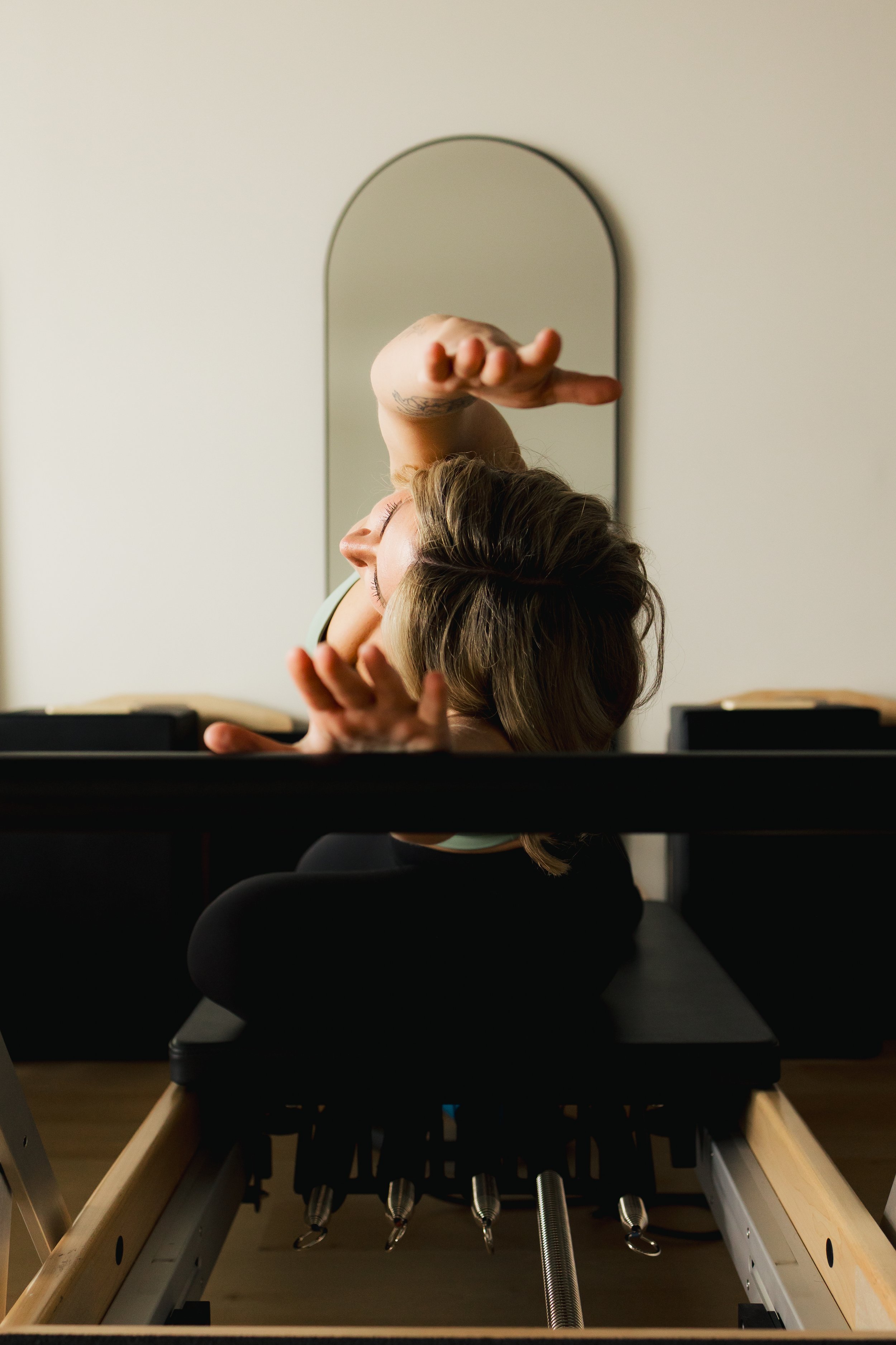 A woman practicing Pilates on a reformer machine, with her arms raised and eyes closed, in a room with a mirror behind her.