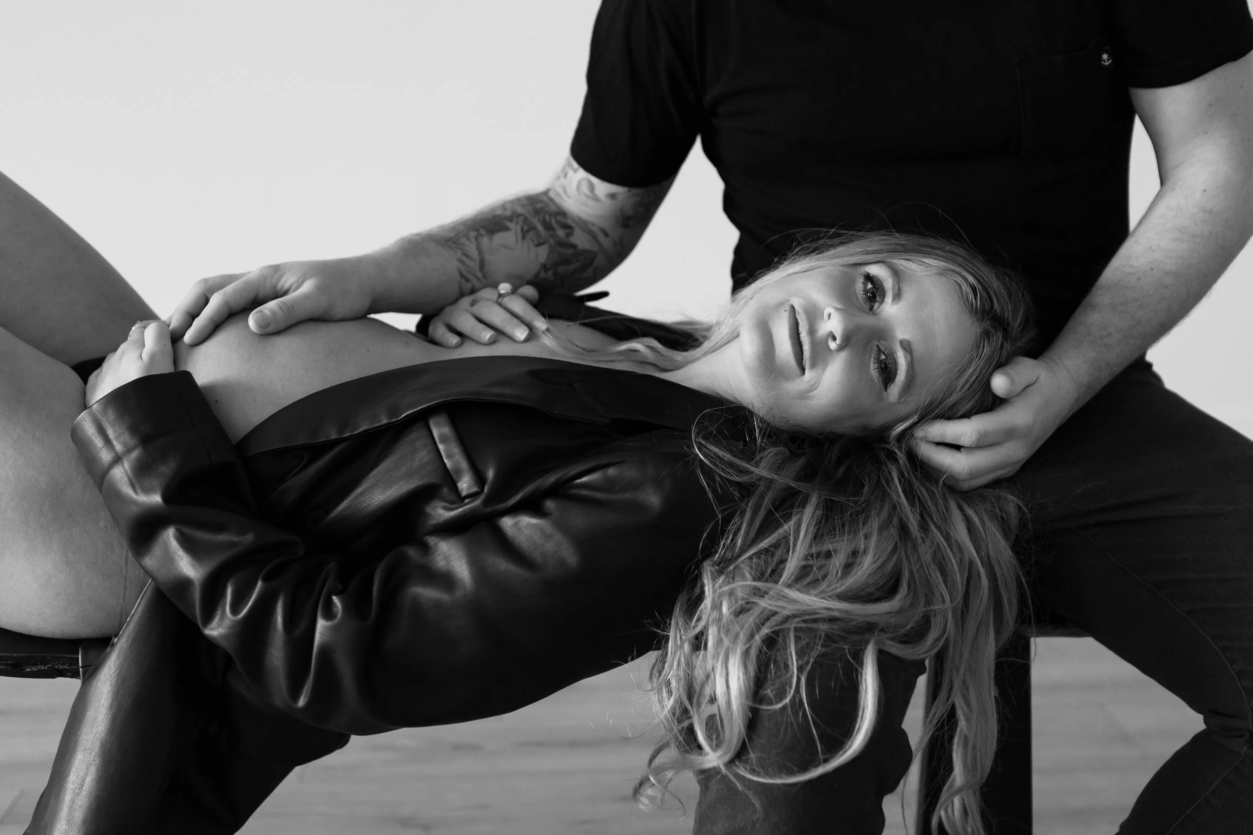 A woman with long wavy hair lying on her side, wearing a leather jacket, with a man providing support by holding her head and arm.
