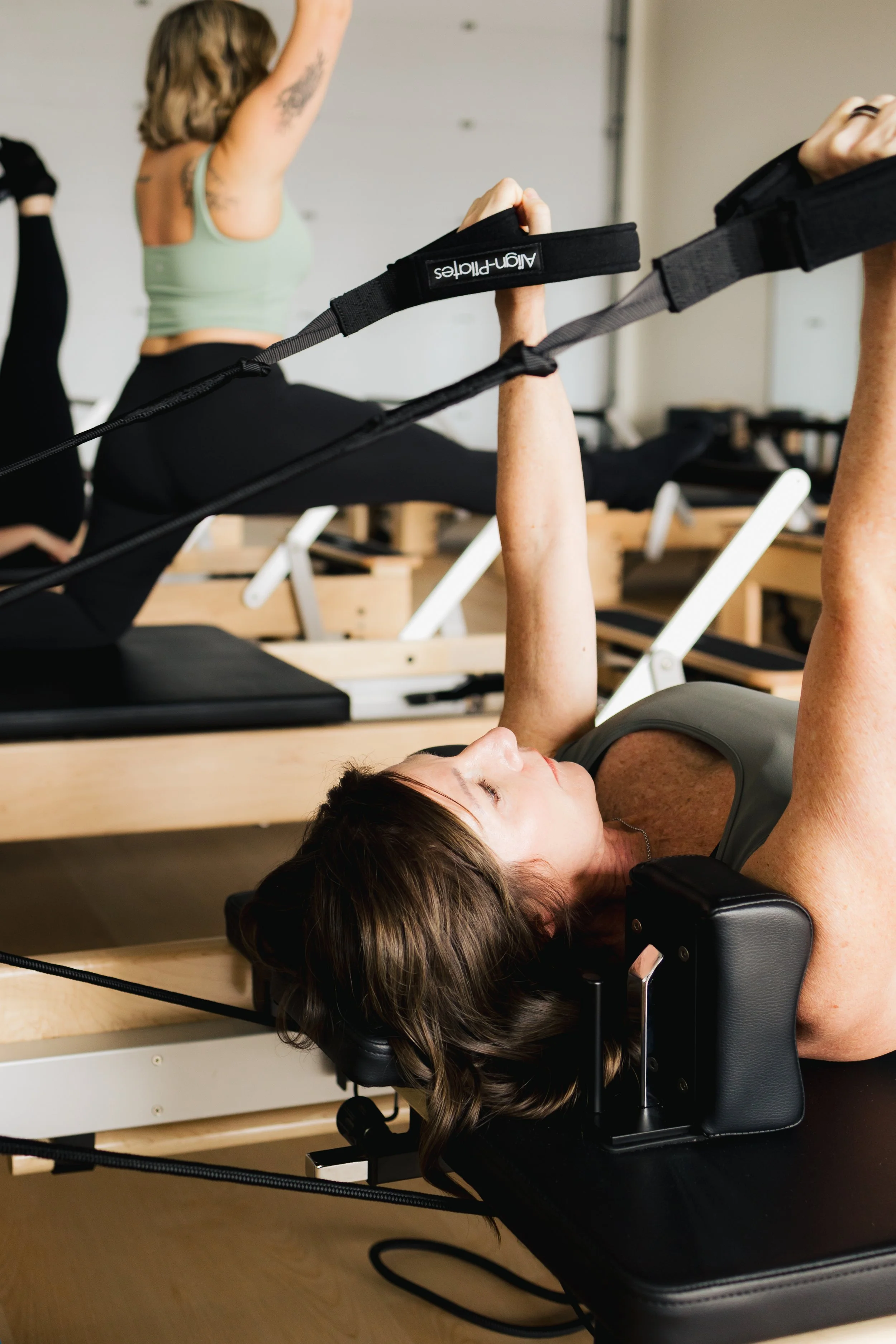 Woman lying on a Pilates reformer machine, holding and pulling on a suspension trainer with her arm extended, in a Pilates or fitness studio with other participants and equipment in the background.