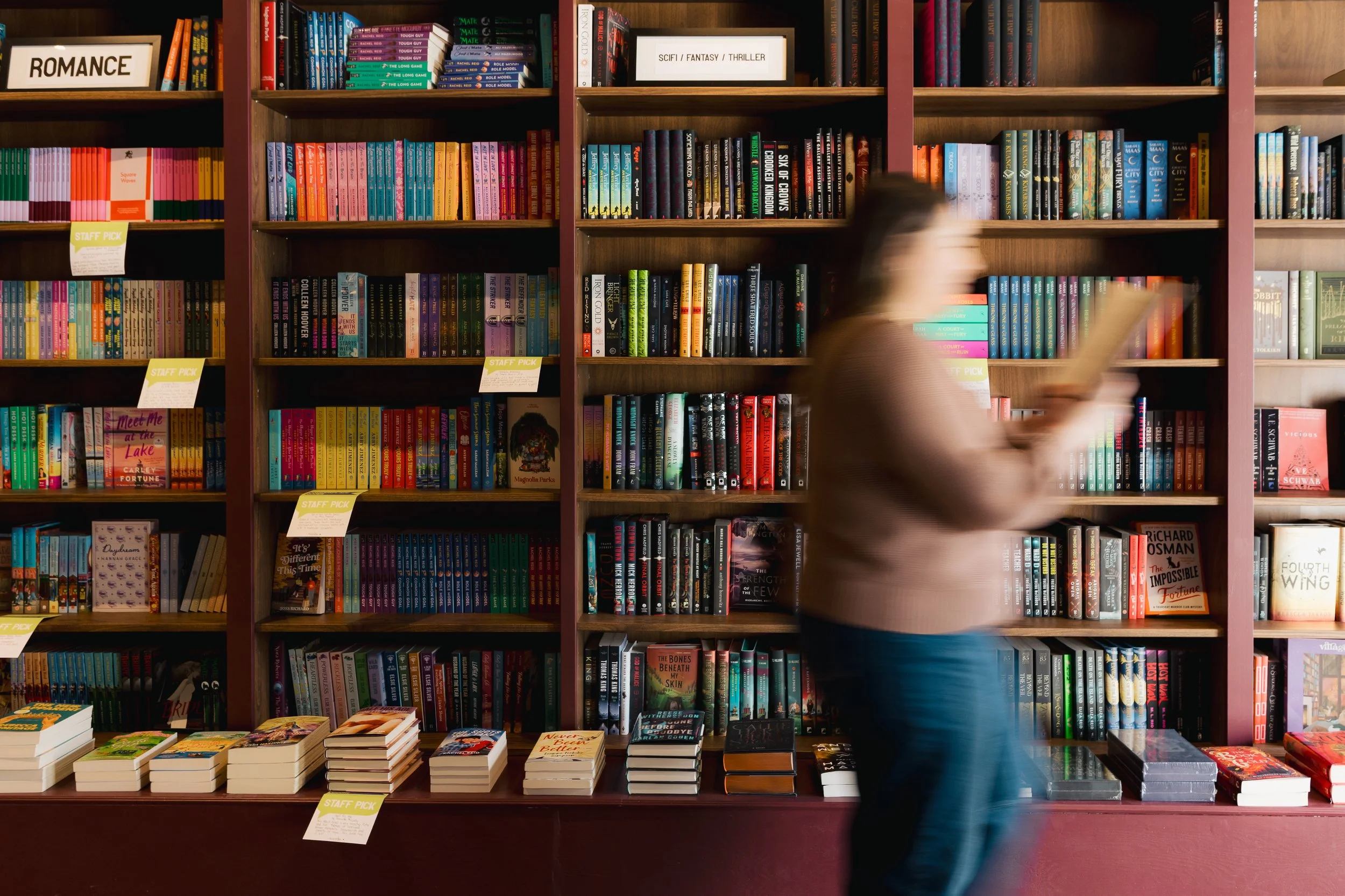 Blurred woman walking past a bookshelf filled with colorful books at a bookstore. Books are arranged on shelves with some displaying yellow 'Staff Pick' tags.