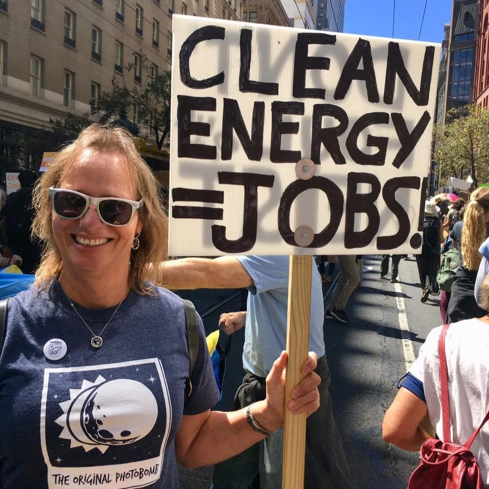 A woman smiling at a climate change protest holds a sign that reads 'Clean Energy = Jobs!'. She is wearing sunglasses, a dark T-shirt with a moon and space design, and has a button pin on her shirt.