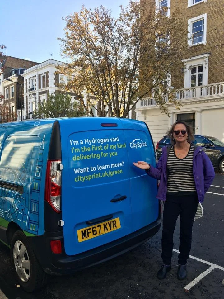 A woman standing next to a blue CitySprint hydrogen delivery van on a city street with trees and buildings in the background.