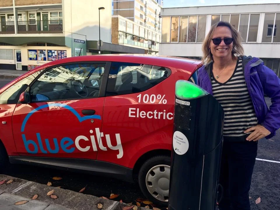 A smiling woman in sunglasses and a striped shirt stands next to an electric car parked on the street. The car is red with the words 'blue city' and '100% Electric' written on it, and it is charging at an electric charging station in an urban area.