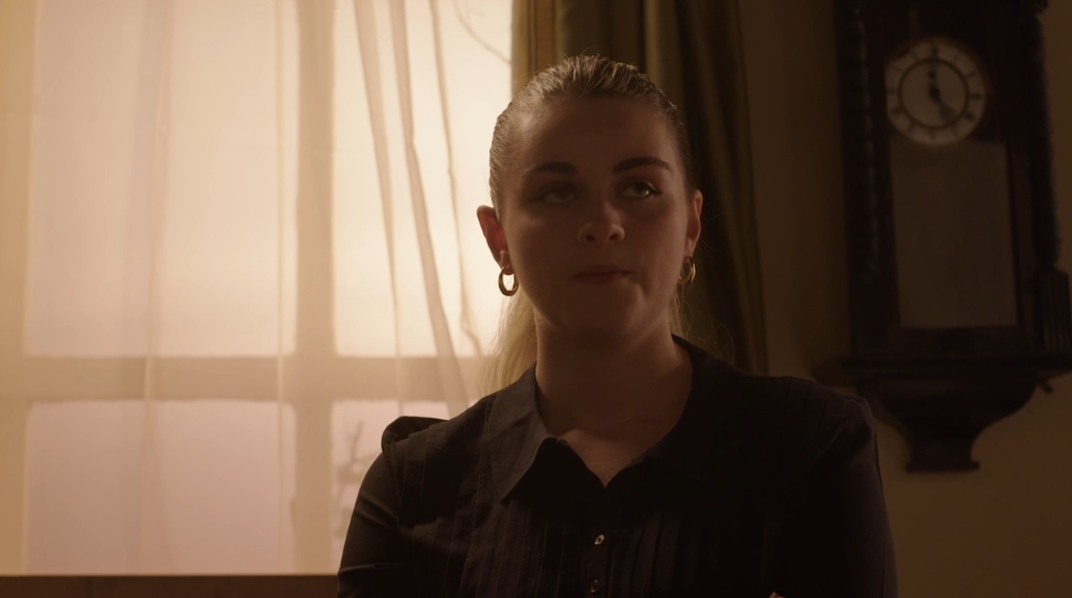 A woman with earrings sitting indoors near a clock, with light filtering through curtains behind her.