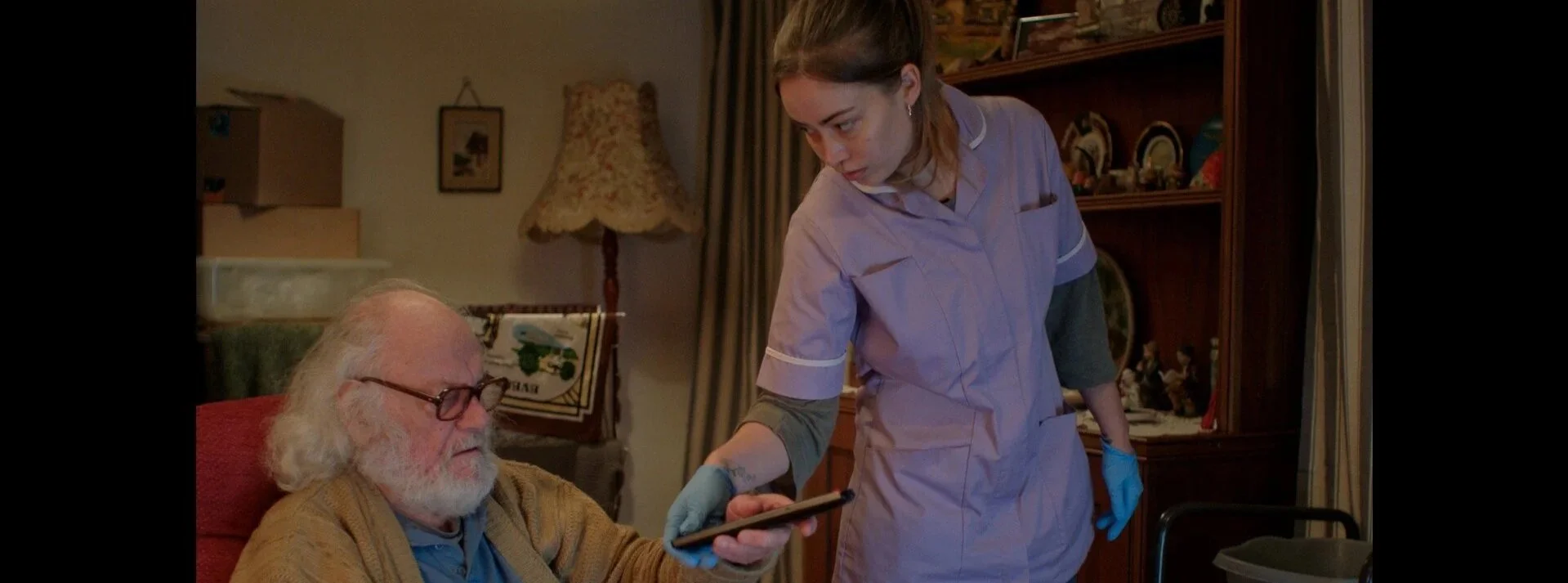 A young woman in pink nursing scrubs and gloves showing something on her phone to an elderly man with white hair and a beard, glasses, who is sitting in a red chair in a warmly decorated room.