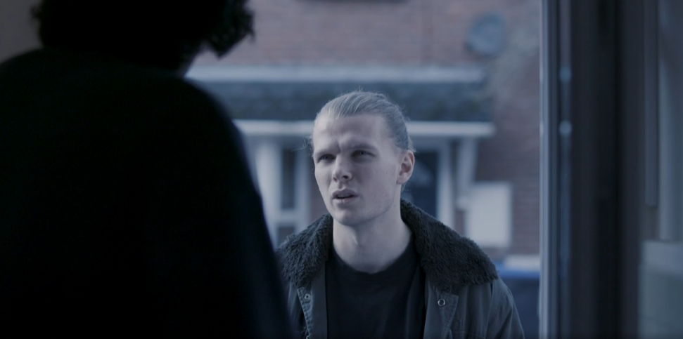 A young man with long blond hair tied back wearing a dark jacket with a fur collar, looking confused or surprised at another person outside.