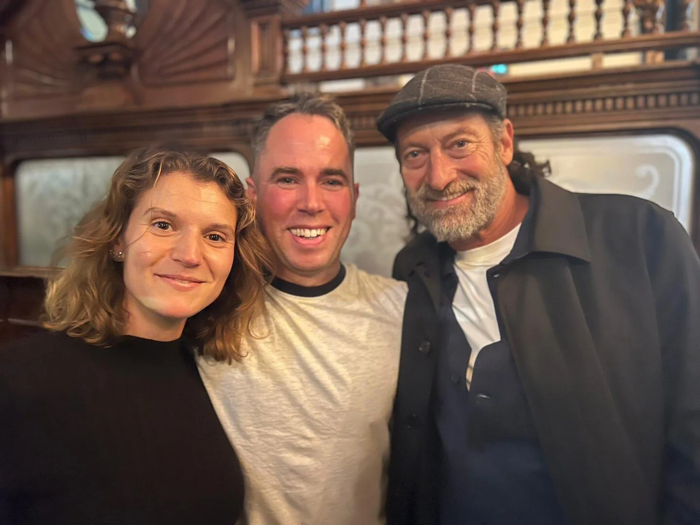 Three people posing together, smiling inside a wooden-paneled room, with a large window in the background.