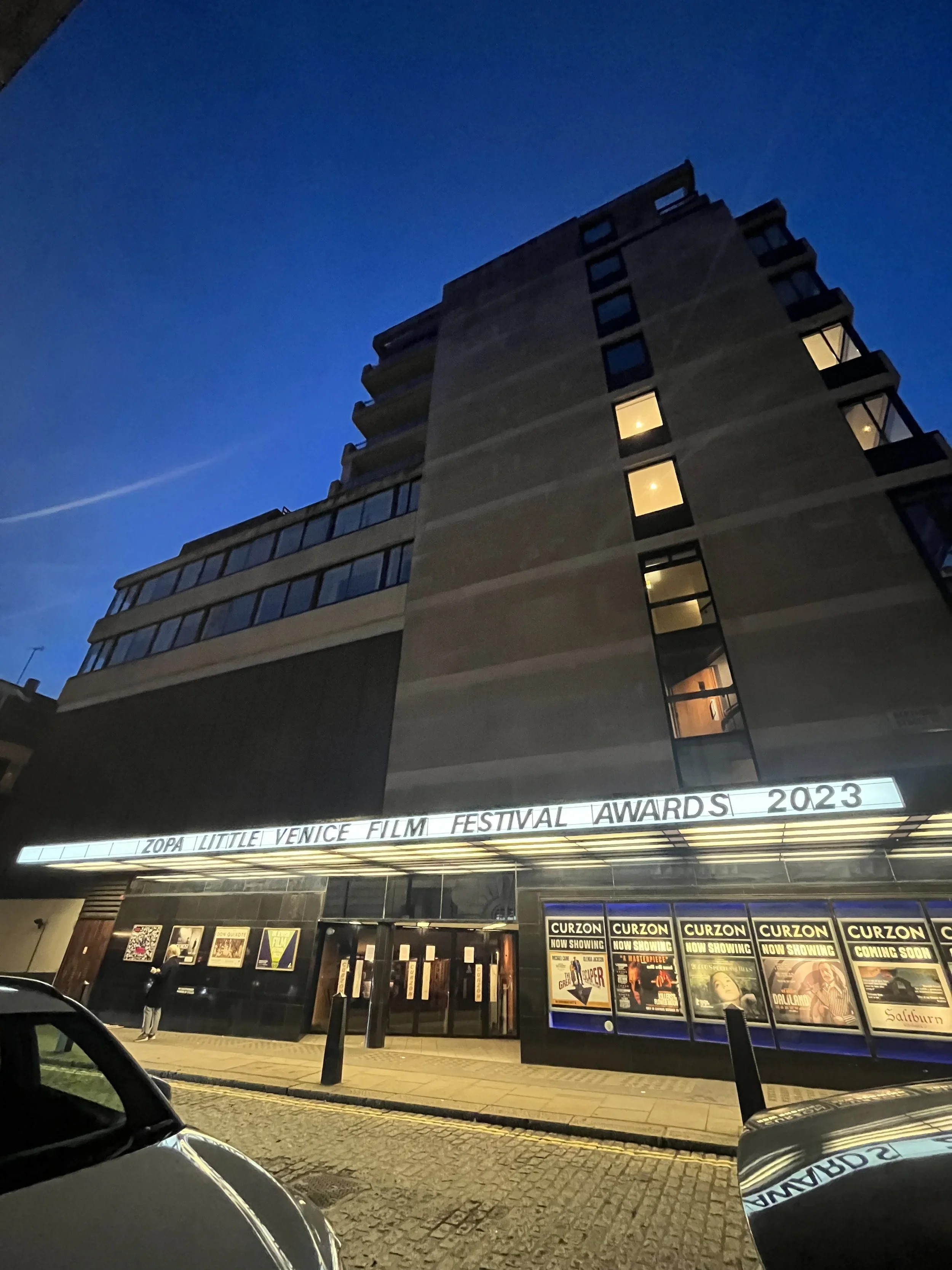 Exterior of a theater building at night with a marquee sign that reads "Venice Film Festival Awards 2023" and posters for upcoming movie screenings.