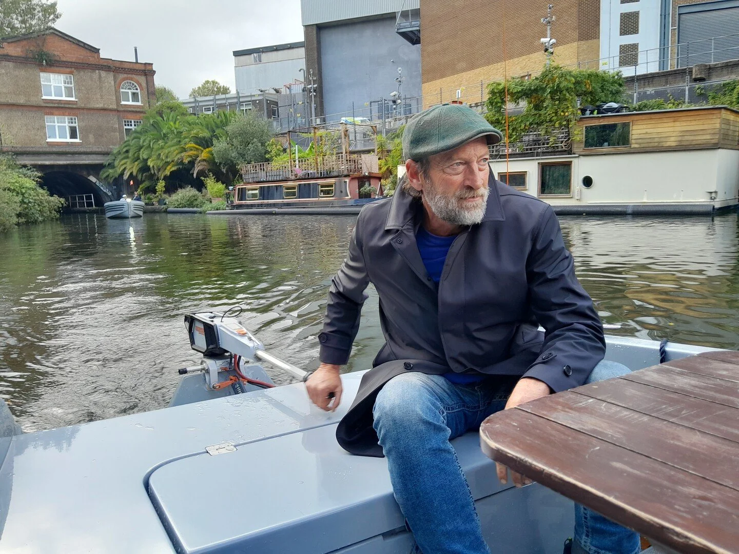 A man with a gray beard and gray hair, wearing a gray cap, navy jacket, and blue jeans, is sitting on a boat by a river, looking to the side. There are houseboats, buildings, trees, and another boat in the background.